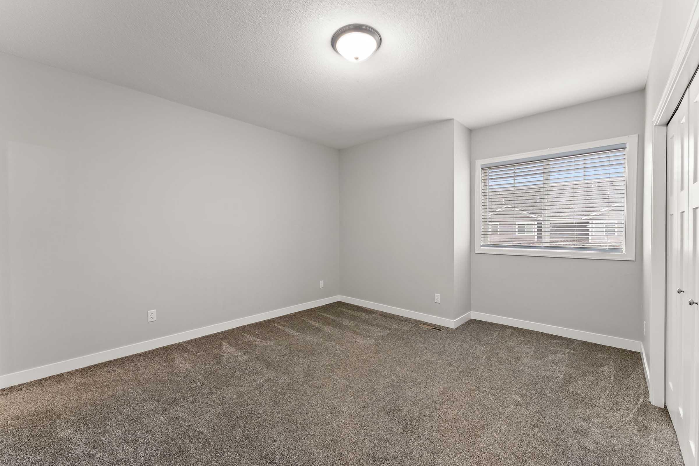 A blank, unfurnished room with light gray walls and soft carpet flooring. The room features a window with closed blinds allowing natural light, and recessed lighting on the ceiling. There is a closet with sliding doors on one side, creating a clean and modern aesthetic.
