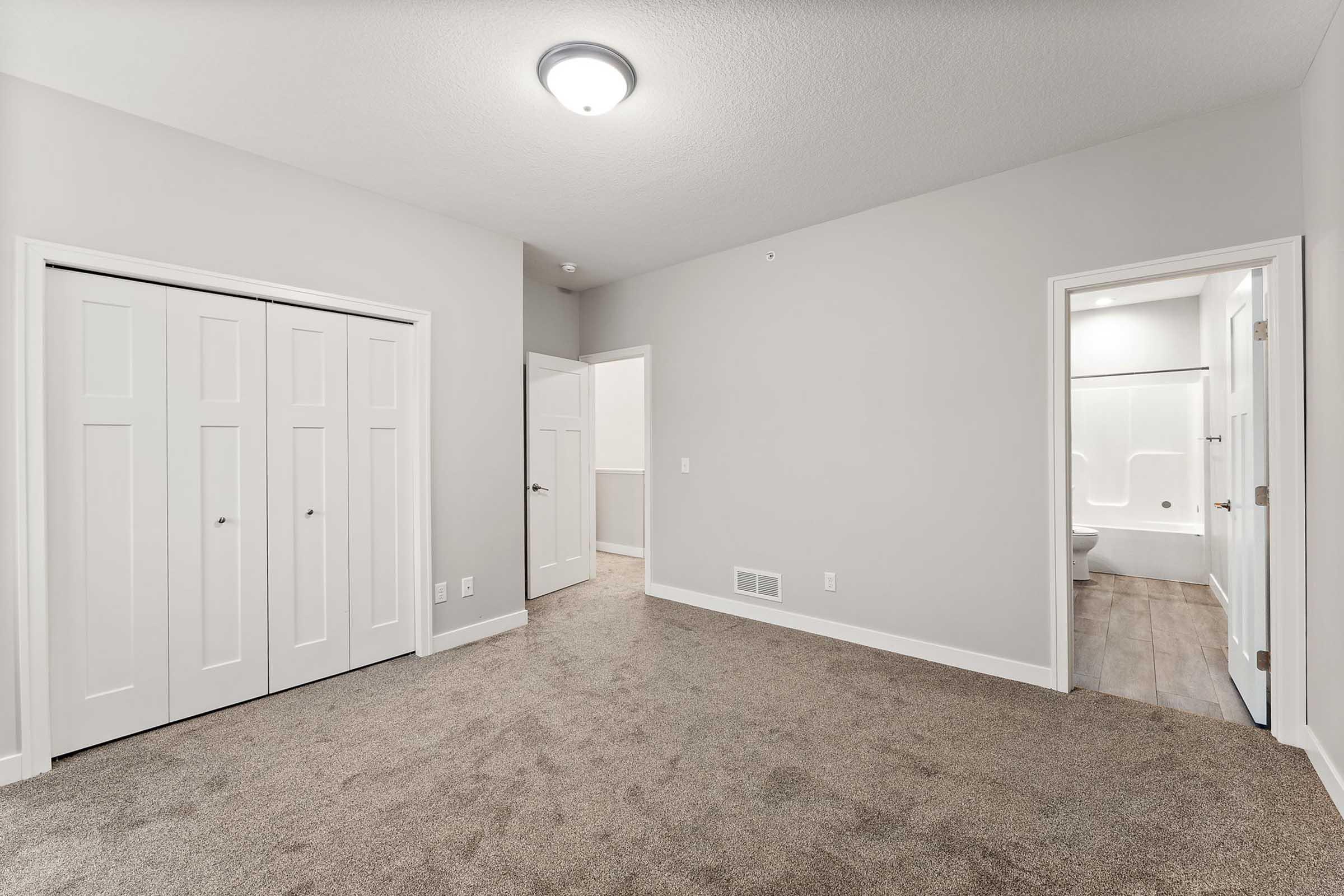 A spacious, light-colored room featuring carpeted flooring, a ceiling light fixture, and two sets of double doors leading to closets. A doorway on the right opens to a bathroom. The walls are painted in a neutral shade, creating a modern and inviting atmosphere.