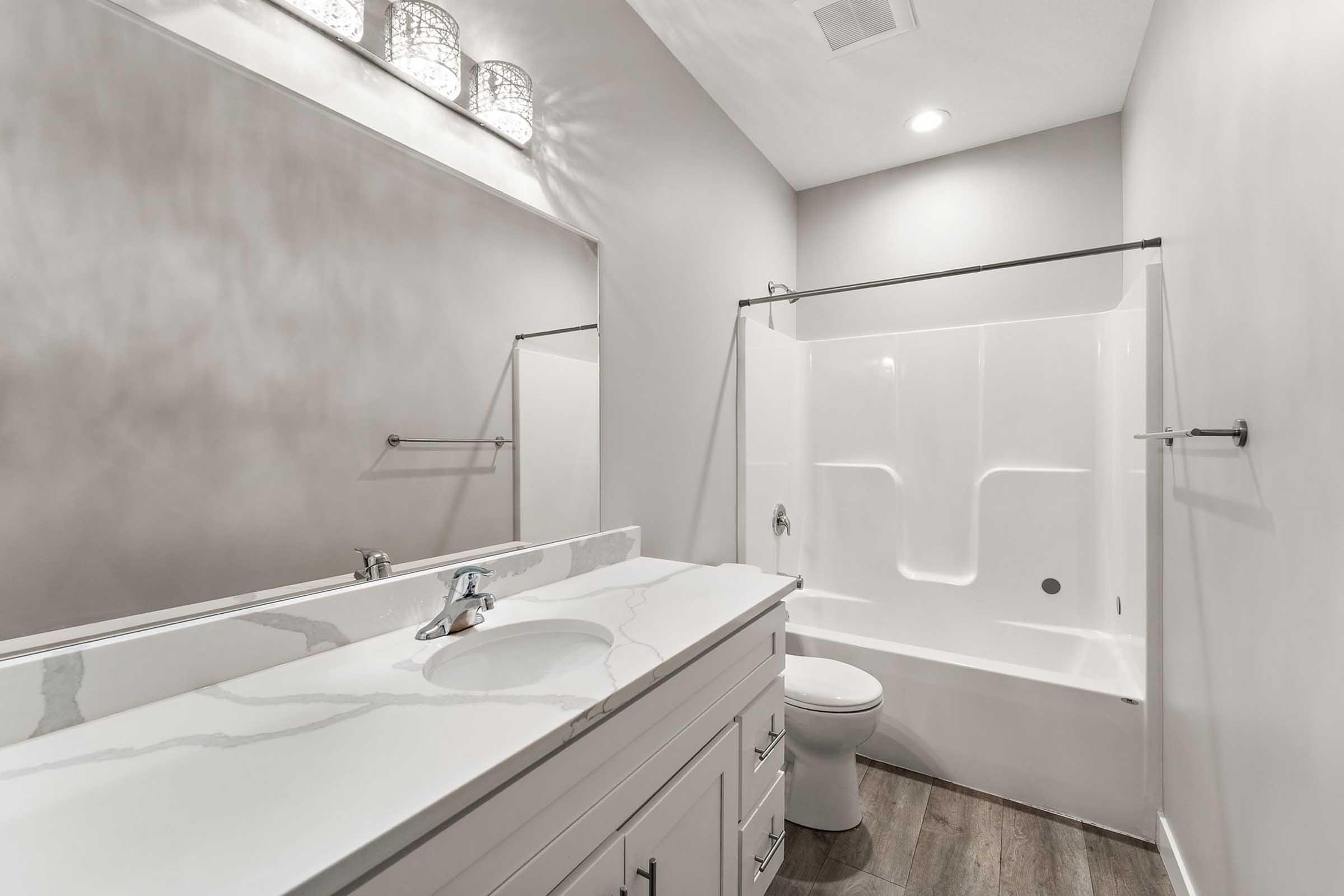 Modern bathroom featuring a sleek white vanity with a marble countertop, a white bathtub with a shower, and a large mirror. The walls are a soft gray, illuminated by recessed lighting and decorative fixtures. The floor is dark wood, creating a contrast with the light colors.