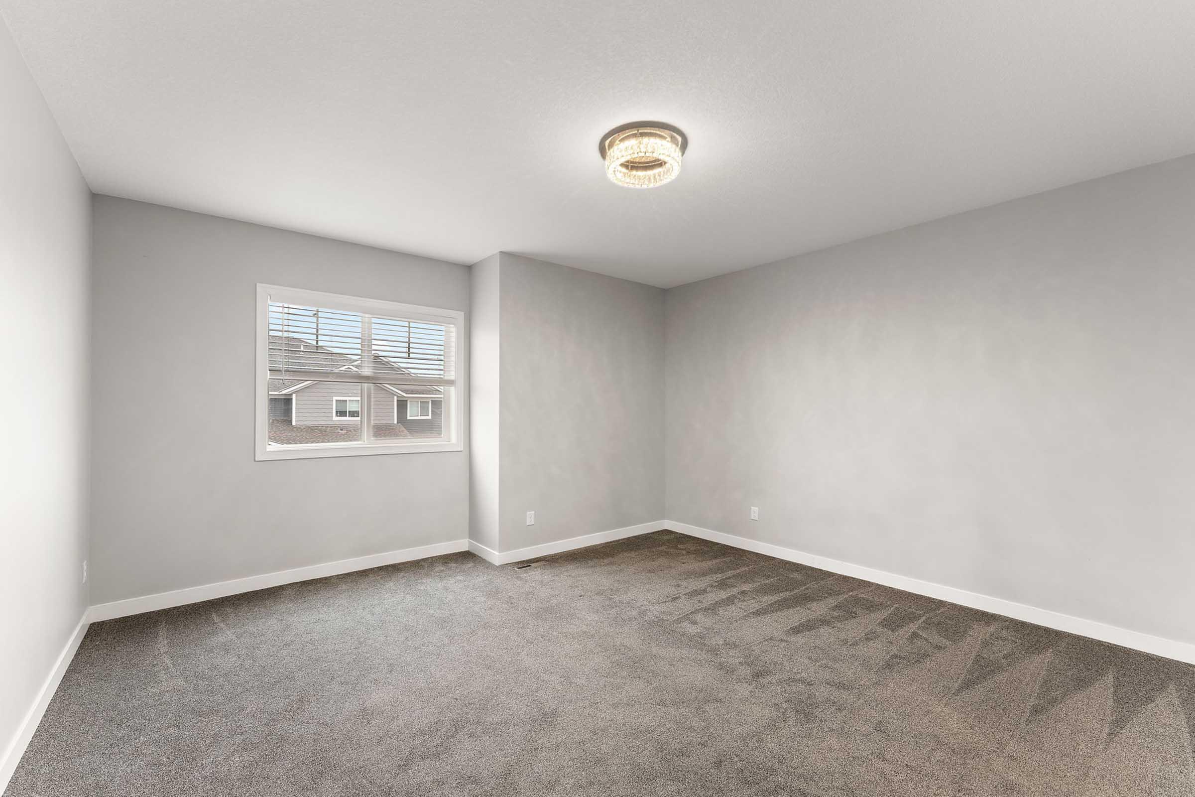 A vacant room with light gray walls and carpeted flooring. It features a modern ceiling light and a window with horizontal blinds, allowing natural light to enter. The room has a minimalist design and is ready for furnishing or decoration.