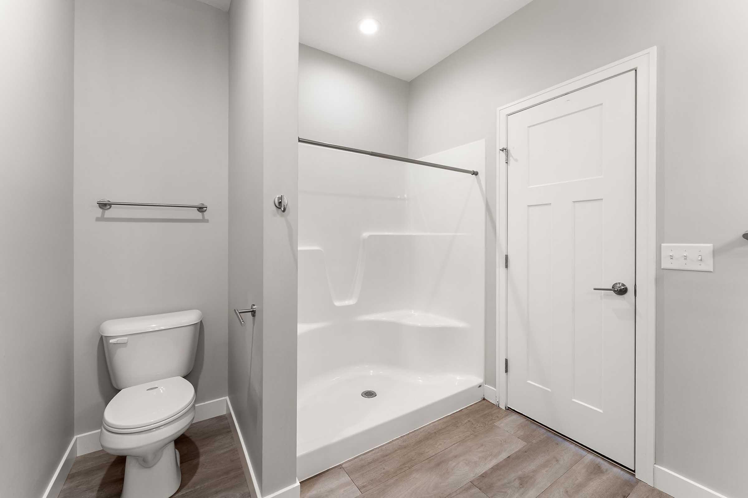 A clean, modern bathroom featuring a shower stall with white walls, a toilet, and a door. The walls are painted light gray, and there is a towel bar mounted on the wall. The flooring is tiled in a neutral color, providing a contemporary and minimalist aesthetic.
