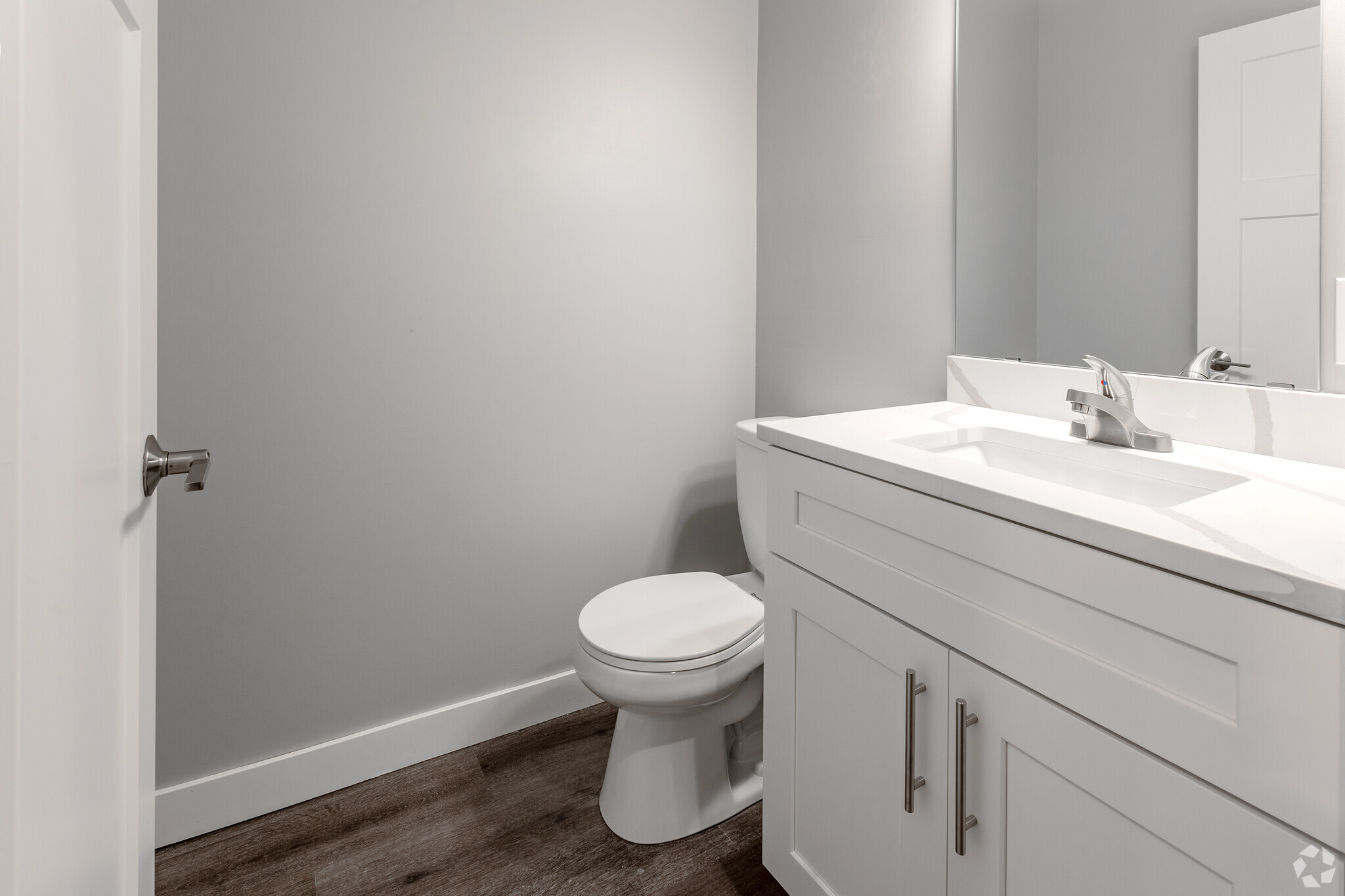 A modern bathroom featuring a white toilet and a sleek vanity with a marble top and sink. The walls are painted light gray, and the flooring is dark wood. A mirror is mounted above the sink, reflecting the minimalist design of the space.