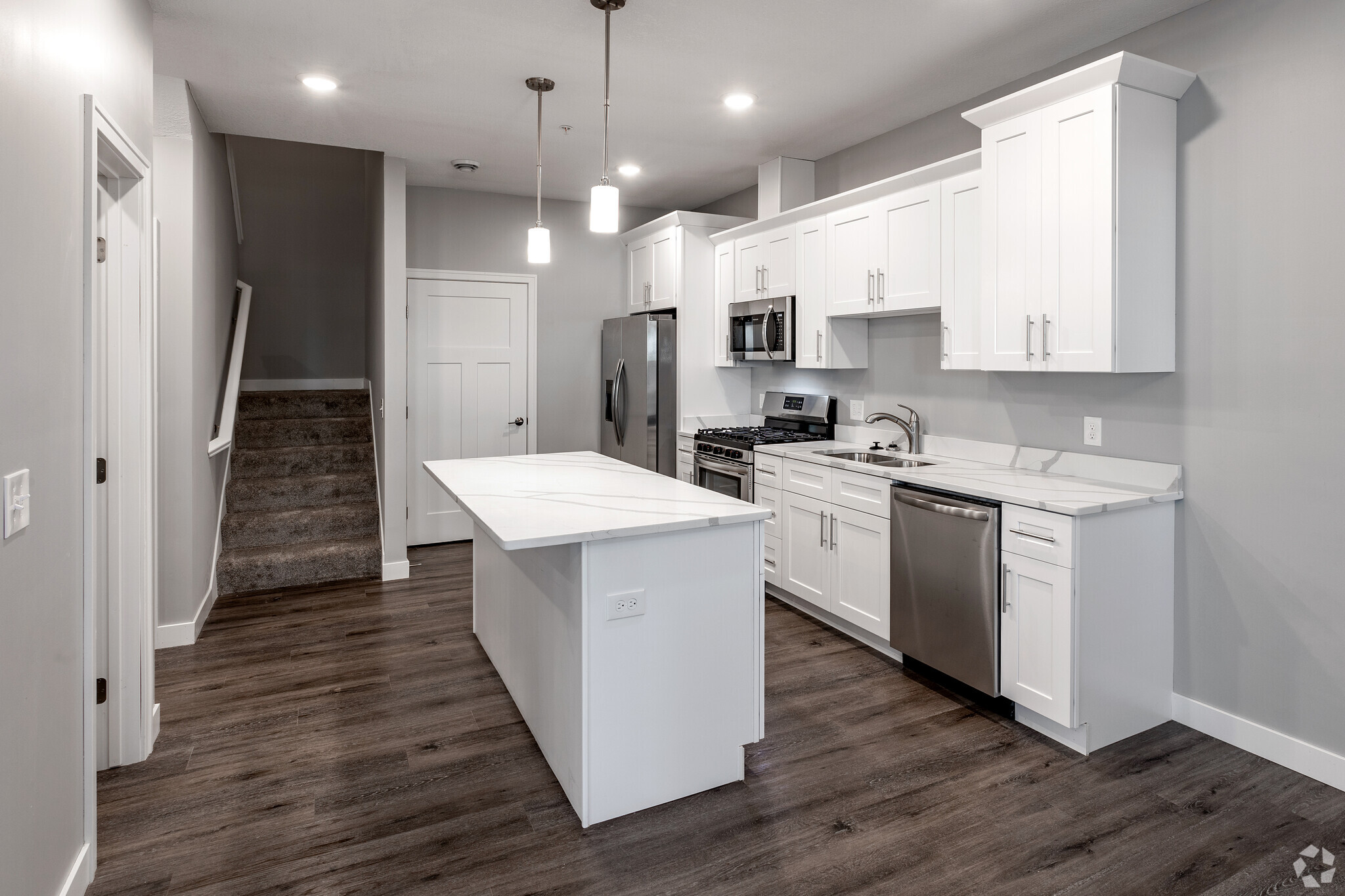 Modern kitchen featuring white cabinetry, stainless steel appliances, and a large island. The open layout includes a staircase in the background, hardwood flooring, and pendant lighting above the countertop, creating a bright and contemporary space.