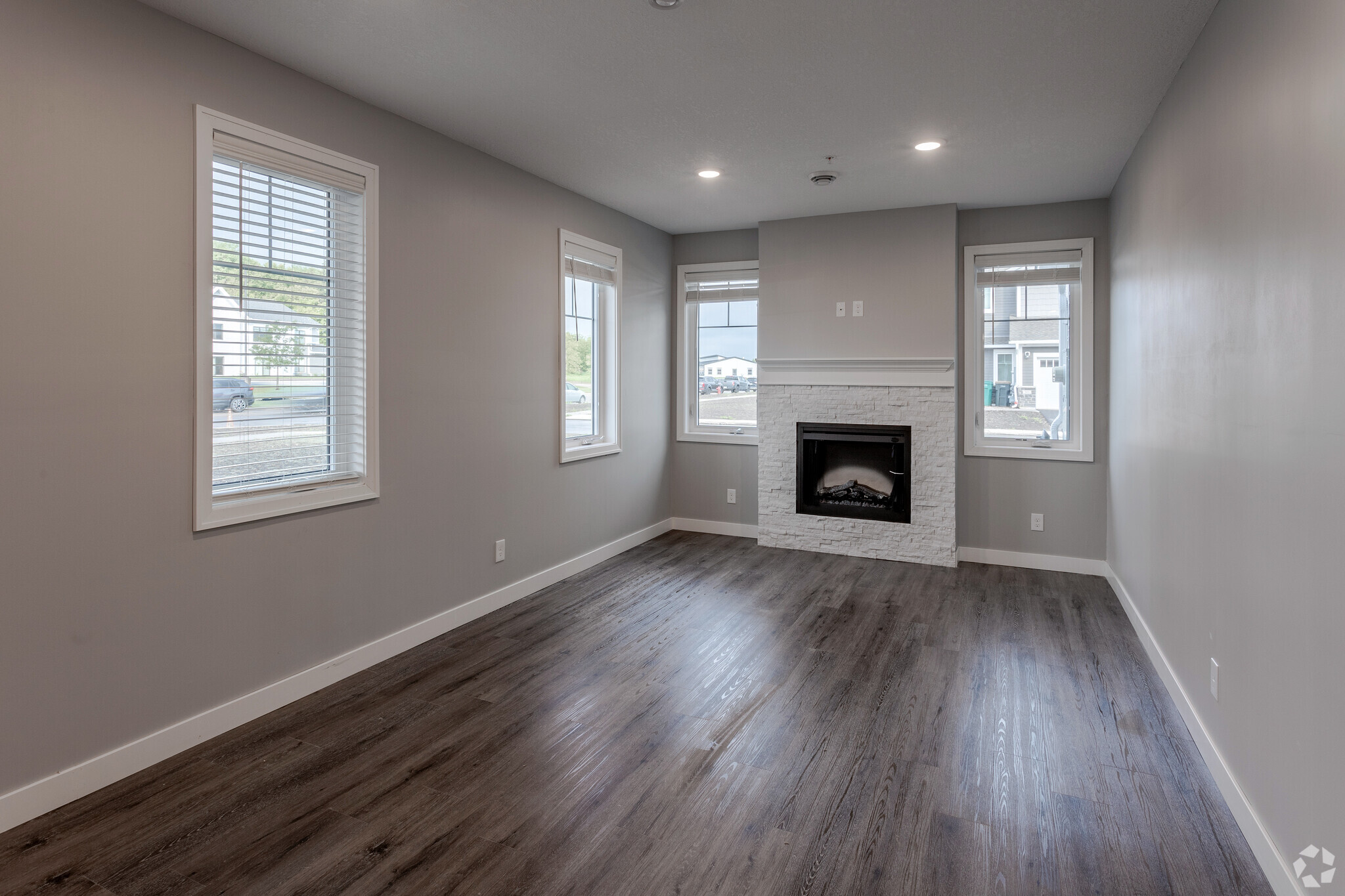 A modern, empty living room featuring hardwood floors, a white stone fireplace, and large windows with horizontal blinds. The walls are painted in a light gray color, creating a bright and airy atmosphere. The room is well-lit, with natural light streaming in from the windows.