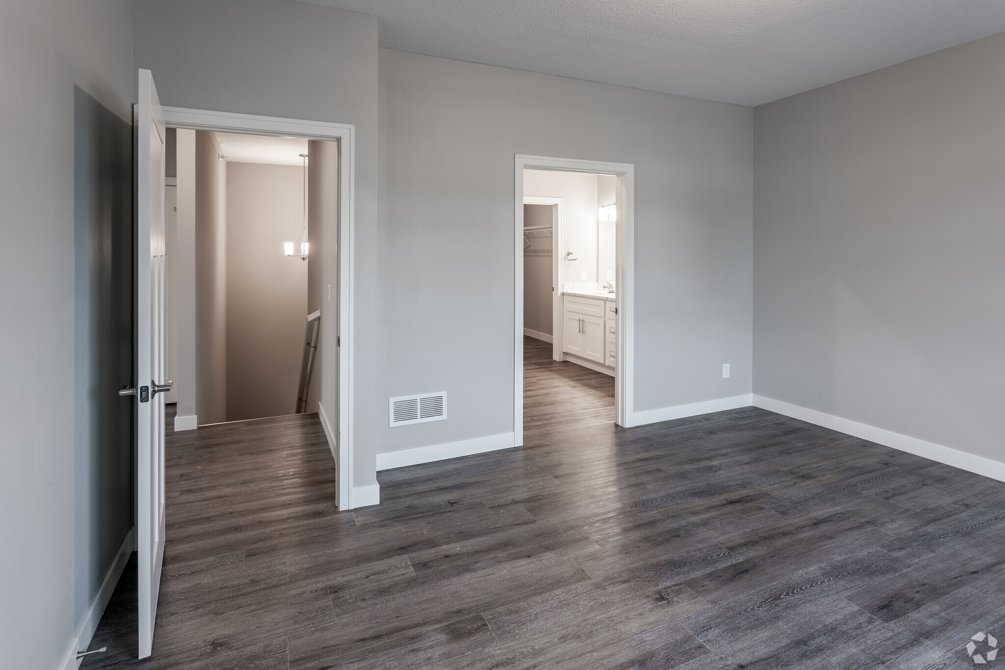 Empty interior of a modern apartment featuring a spacious room with gray walls and dark wood flooring. There are two open doorways: one leading to a bathroom area with a double vanity, and another leading to a staircase. The lighting is bright and neutral.