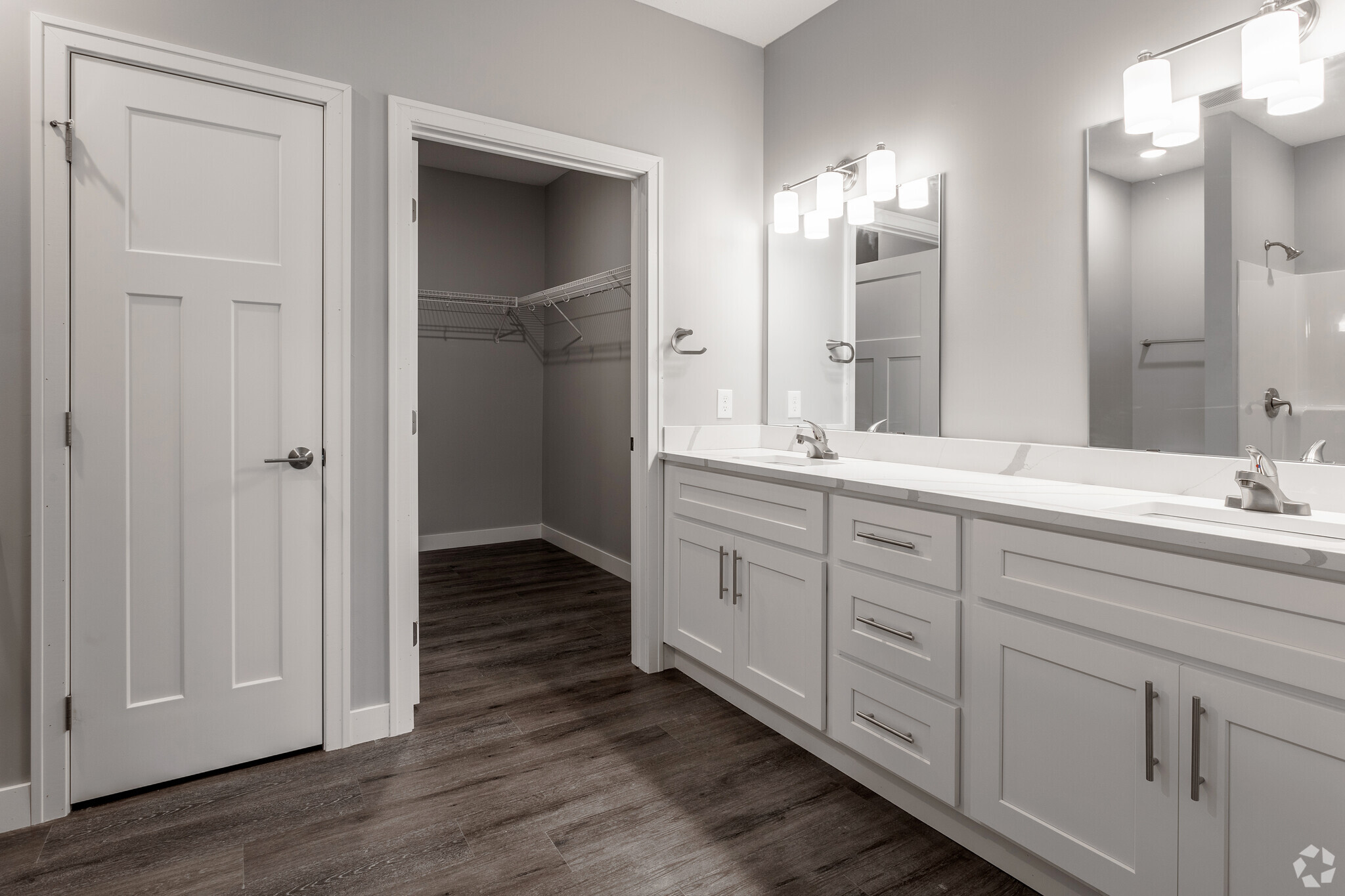 Modern bathroom featuring dual sinks with stylish fixtures, large mirrors above each sink, and a door leading to a walk-in closet. The walls are painted a light gray, and the flooring is a dark wood. The overall design is sleek and contemporary, emphasizing functionality and aesthetics.