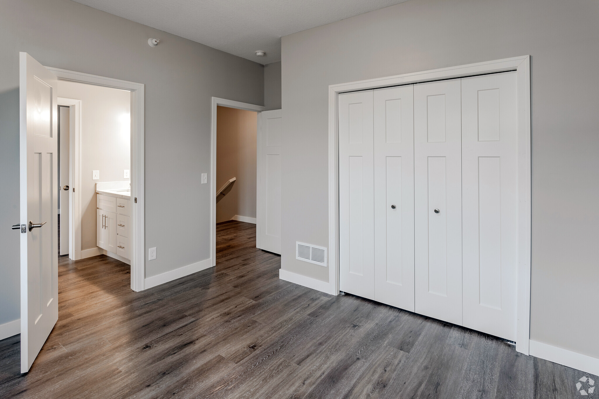 A modern room featuring light gray walls and a wooden floor. Two white doors lead to separate spaces: one to a bathroom with a vanity and the other an entrance to a hallway. A large white closet with multiple panels is visible on the right side of the image. Natural light illuminates the area.