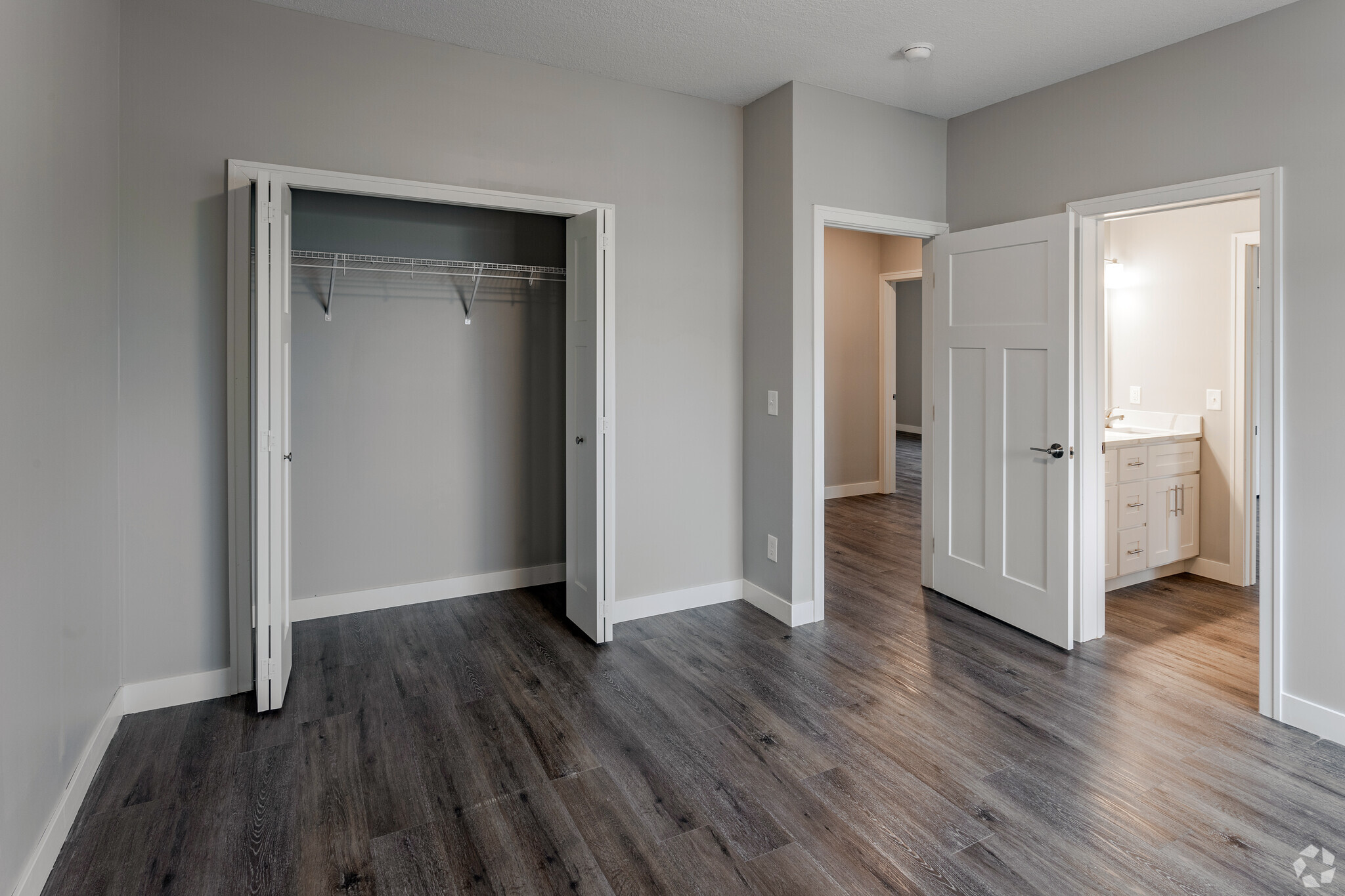 A spacious, empty room with light gray walls and dark wooden flooring. Two open doors lead to a closet and a bathroom. The closet features a wire shelf, and the bathroom doorway is framed with light. Natural light comes from an unseen source, enhancing the room's inviting atmosphere.