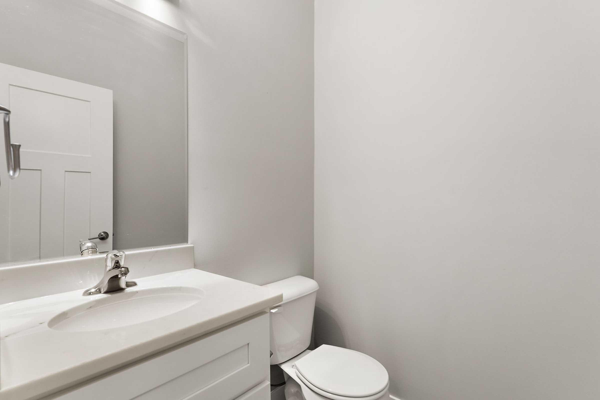 A clean, modern bathroom featuring a white sink with a silver faucet, a white toilet, and plain gray walls. The space is bright and minimalistic, with a mirror above the sink and a door visible in the background.