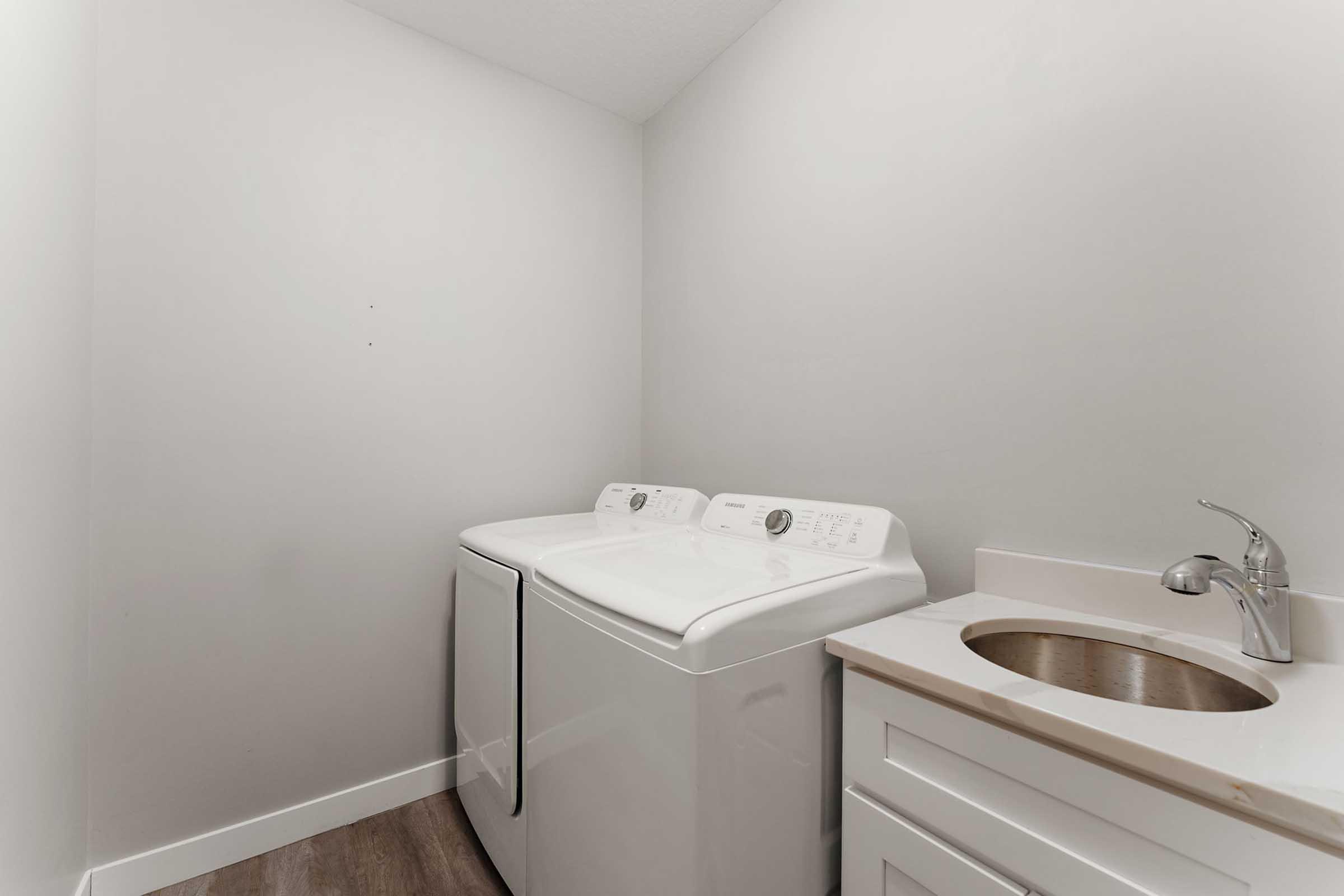 A clean and well-lit laundry room featuring a white washer and dryer against a light gray wall. There's a small sink with a faucet and a white countertop beneath it, along with light-colored cabinetry. The floor is a wood-like laminate, creating a modern and functional space.