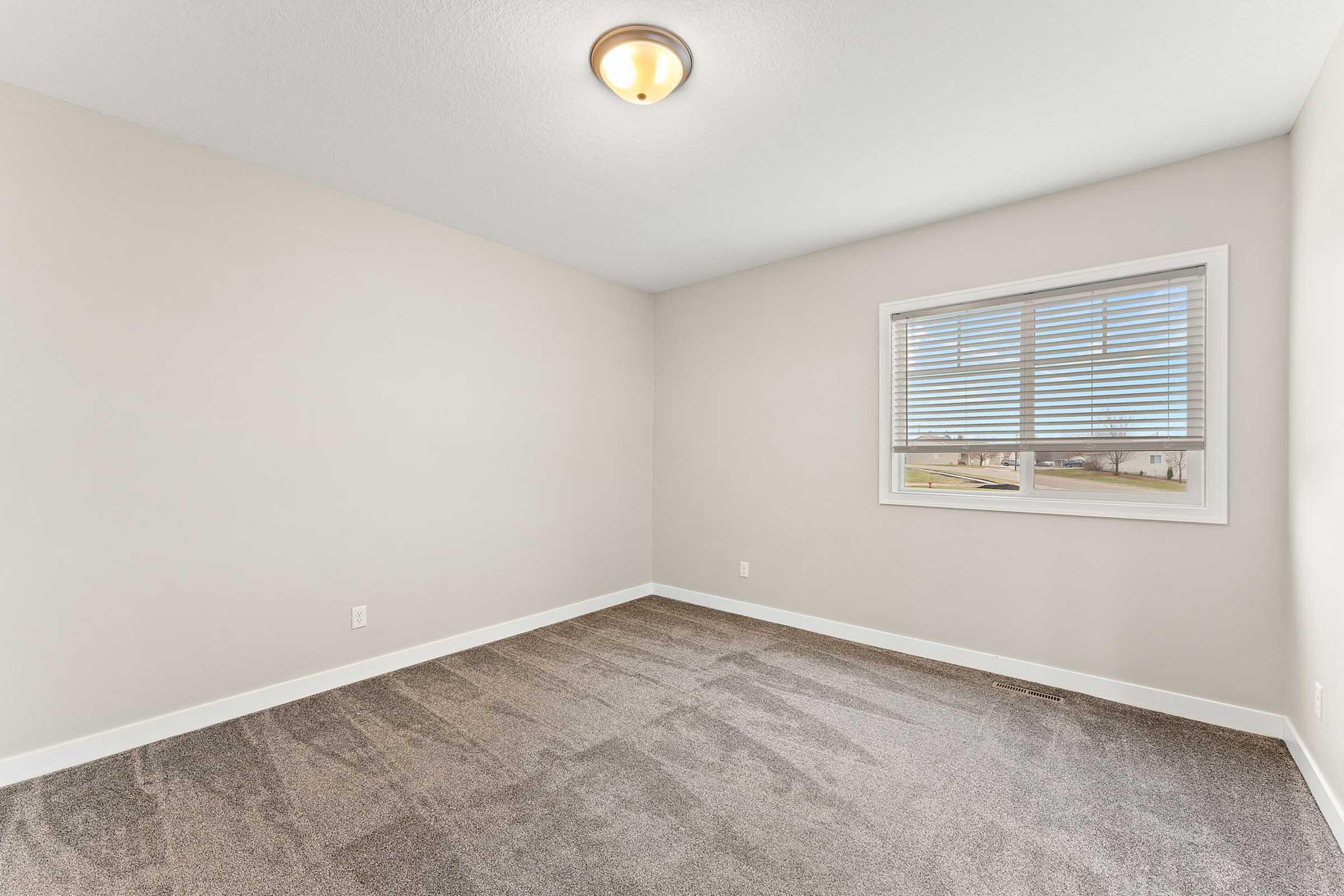 A spacious empty room with light gray walls and a soft carpeted floor. A window with blinds allows natural light to enter, illuminating the space. The ceiling features a simple light fixture, enhancing the clean and modern aesthetic of the room.