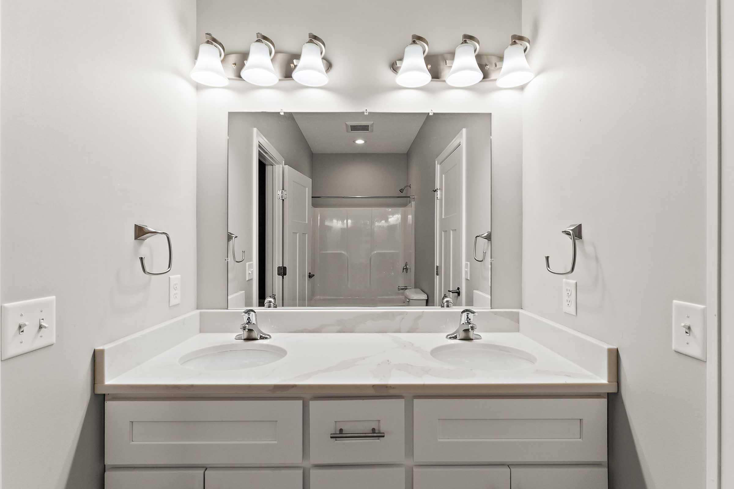 A modern bathroom with a double vanity. The vanity features a white countertop with two faucets and mirrors above. There's adequate lighting from five sconces. In the background, a shower area is visible behind a glass door, and the walls are painted in a light gray color.