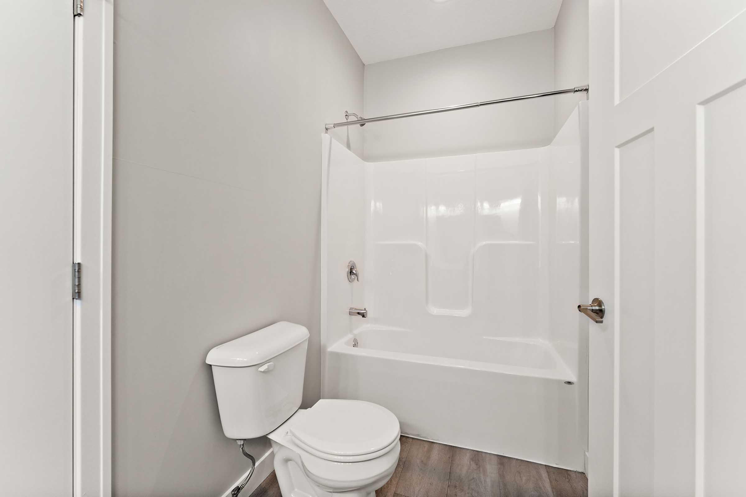 A clean and modern bathroom featuring a white bathtub with a shower curtain rod, a white toilet, and light gray walls. The space has a minimalistic design with wooden flooring and a bright, well-lit atmosphere.
