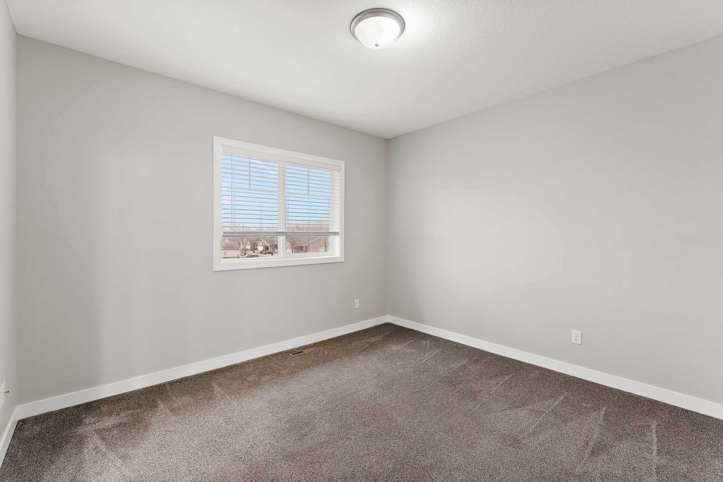 Empty room with light gray walls and a window with blinds. The floor is covered in gray carpet. A ceiling light fixture is visible. Natural light enters through the window, illuminating the space. The room appears clean and uncluttered, ideal for personal customization.