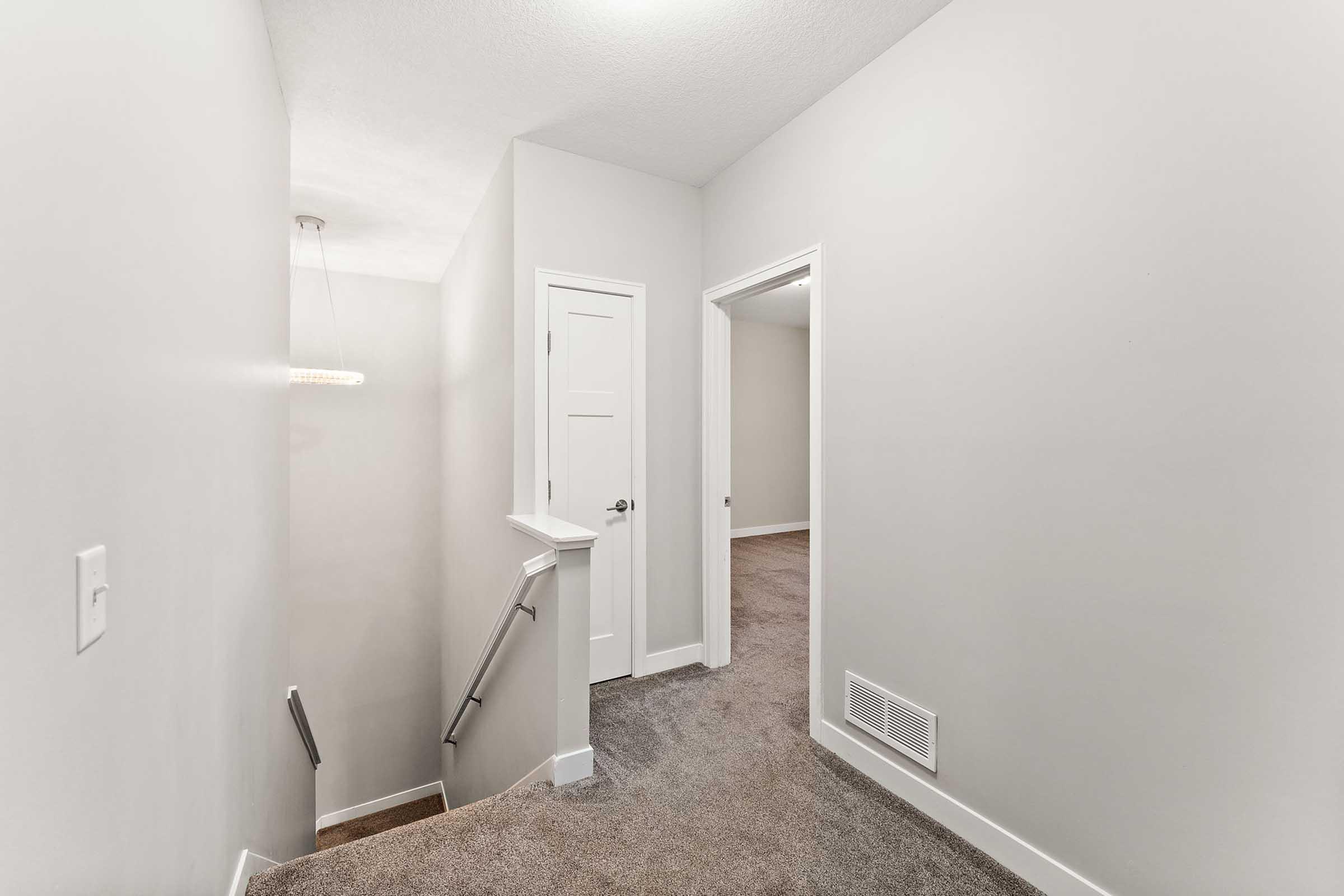 A well-lit, neutral-colored hallway featuring carpeted stairs, leading to two doorways: one to a room and another closed door. A modern ceiling light fixture hangs above, and the walls are painted in a light gray tone. The space appears clean and inviting.
