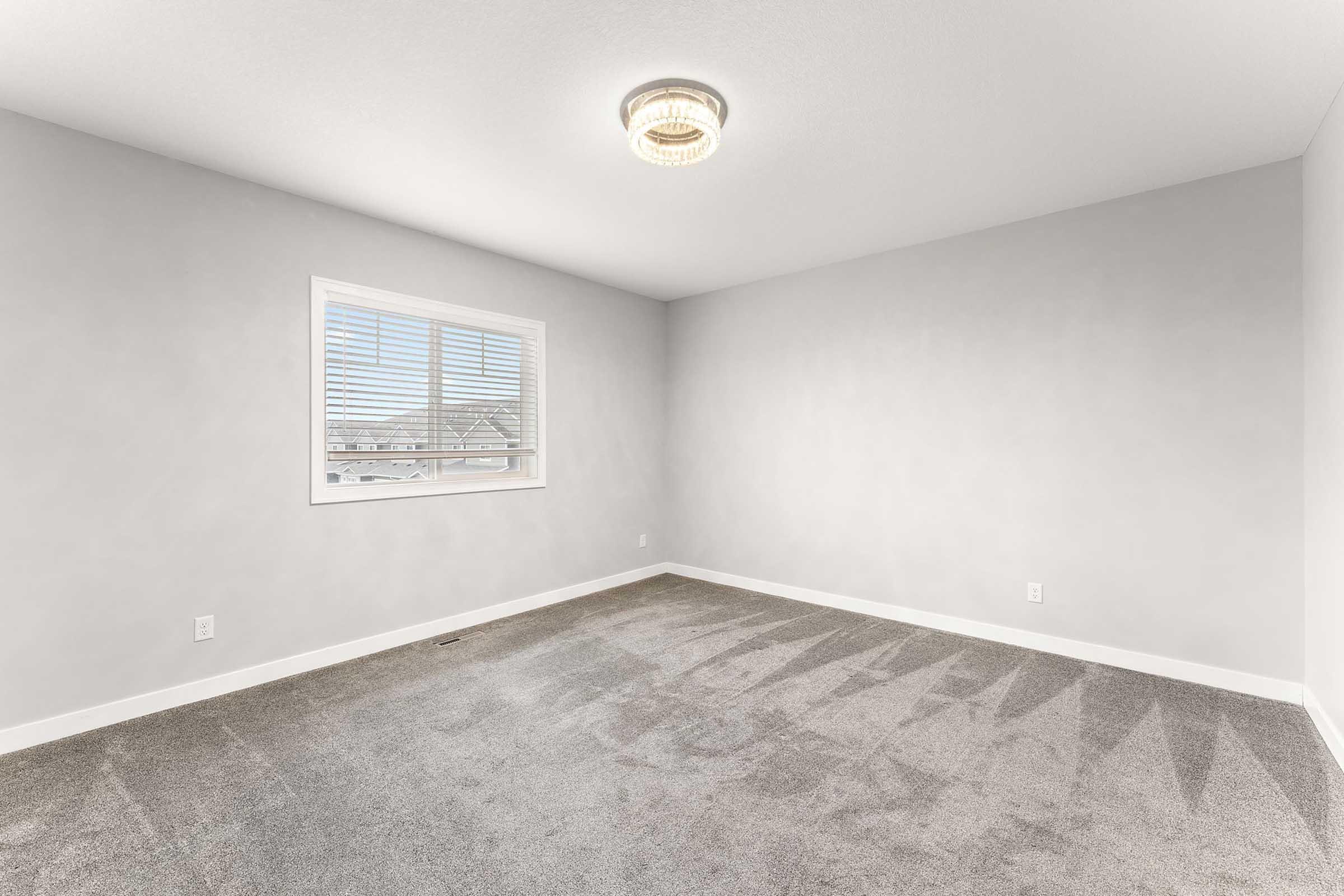 A vacant room with a light gray painted wall and light gray carpet. A window with white blinds allows natural light to enter, casting shadows on the carpet. The room features a modern ceiling light fixture and has no visible furniture or decorations.