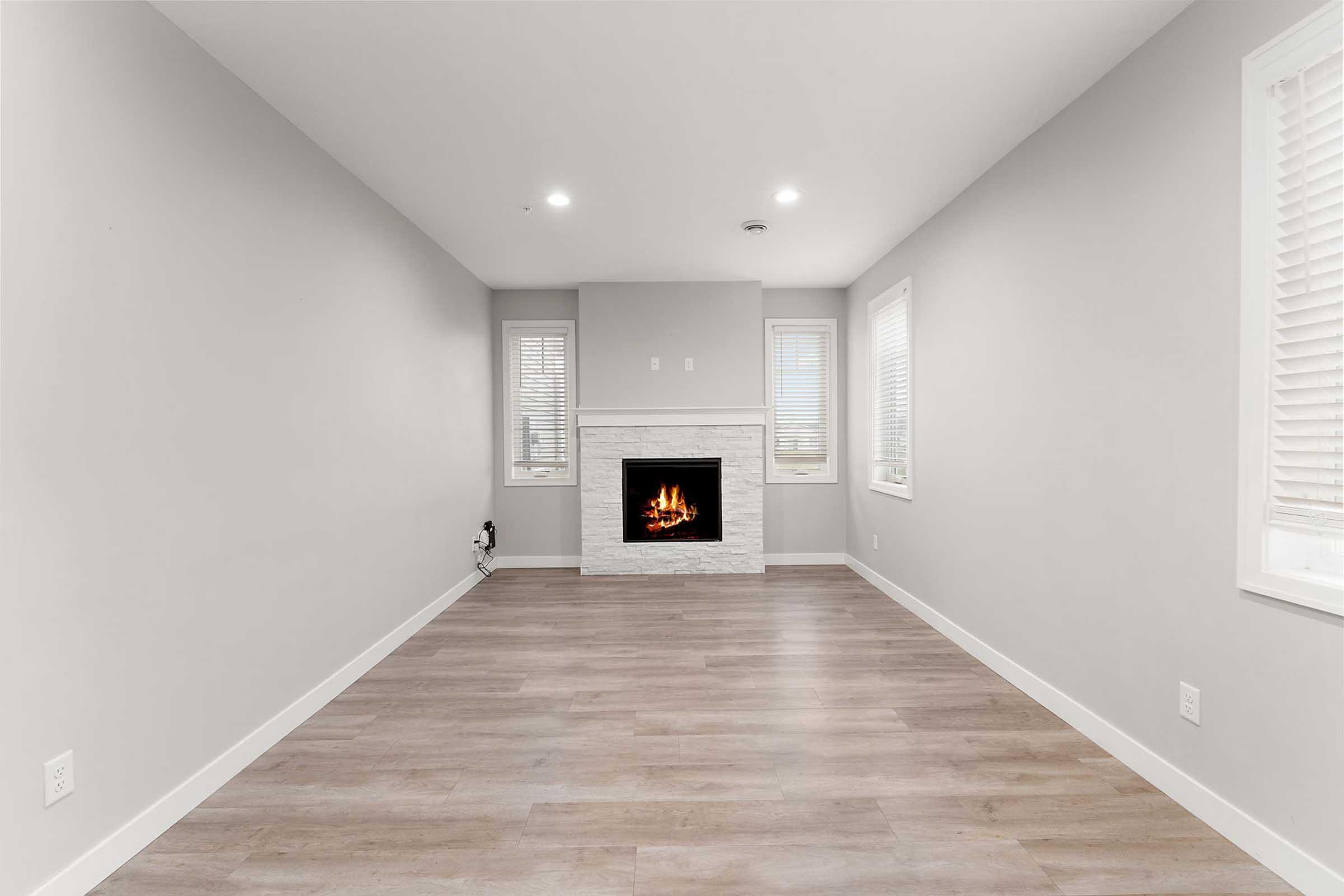 A minimalist interior of a modern living room featuring light gray walls, a white stone fireplace at the center, and two windows with blinds on either side, allowing natural light to enter. The flooring is light wood, and the space is uncluttered and ready for furniture.
