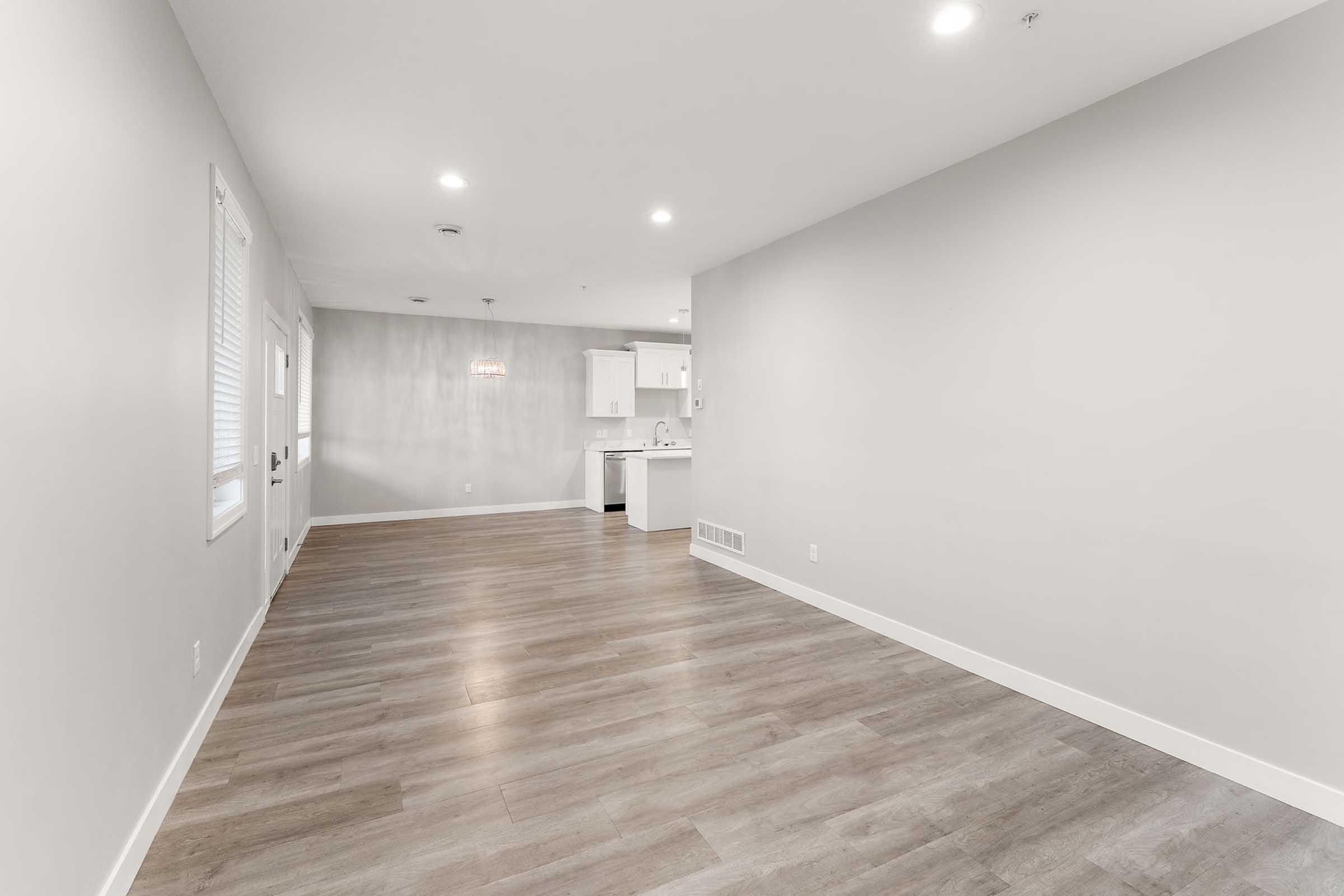 A spacious, empty room featuring light-colored walls and modern wooden flooring. Natural light enters through a window, and a kitchen area is visible in the background with white cabinetry. The space is bright and clean, inviting for various interior design possibilities.