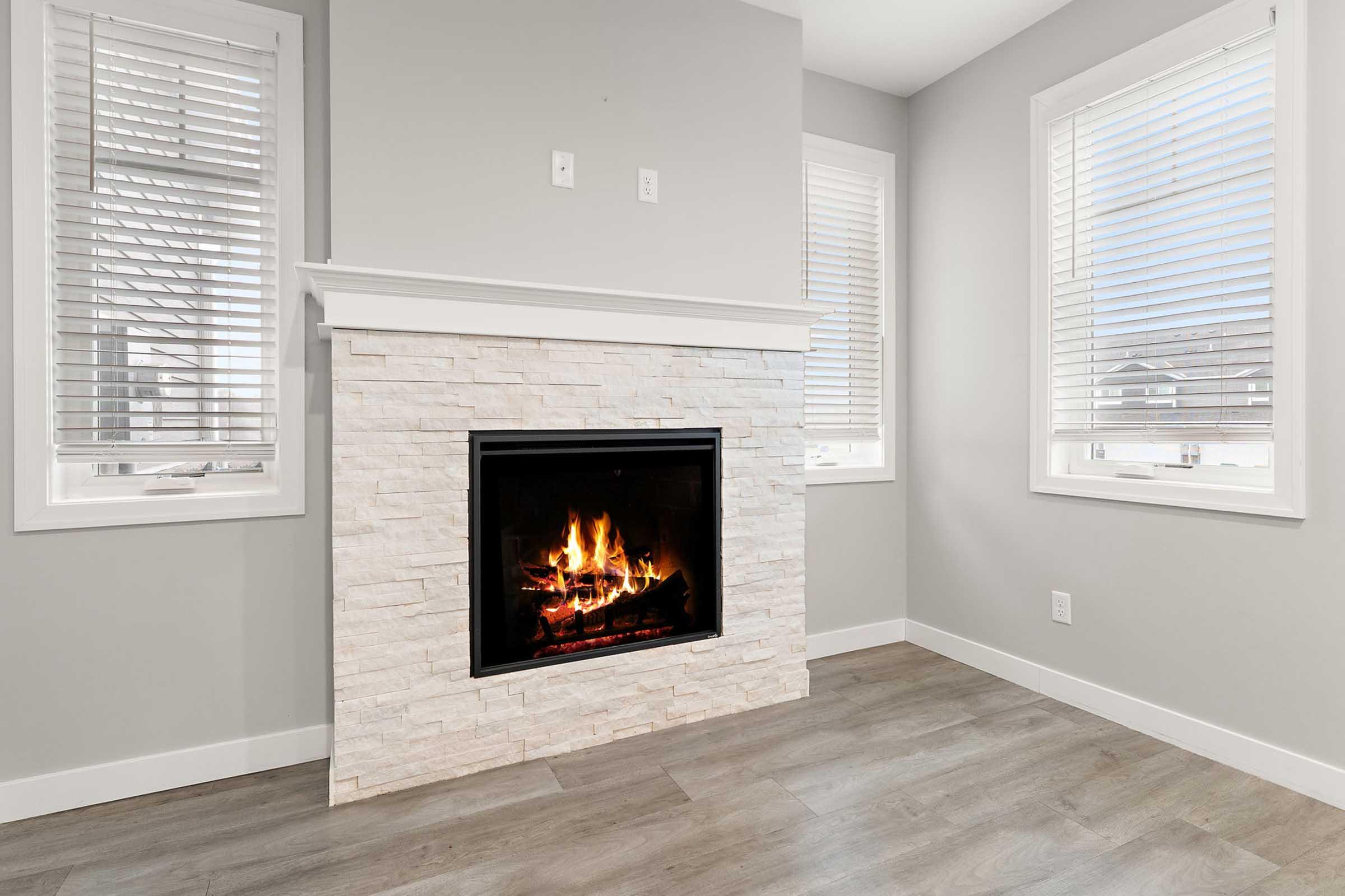 A cozy living room featuring a modern fireplace with a decorative stone facade. The room has large windows with blinds, allowing natural light to flood in. The walls are painted light gray, and the flooring is a warm wood-look finish, creating a welcoming atmosphere.