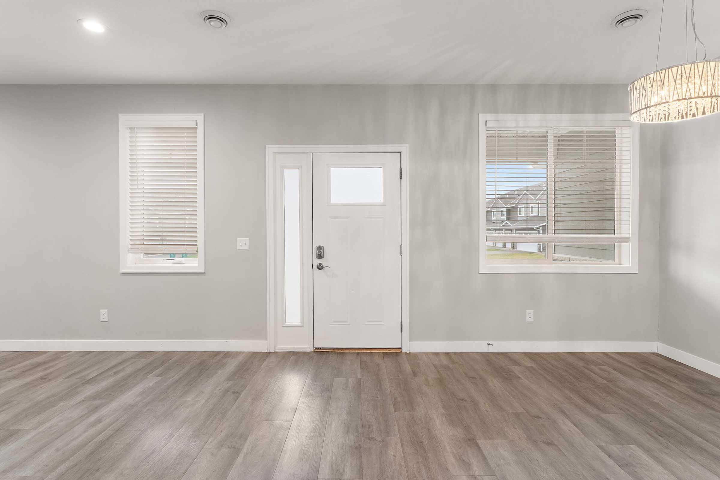 Empty interior of a modern room with light gray walls and wooden flooring. There's a white front door with a window, flanked by two windows with blinds. A circular chandelier hangs from the ceiling, adding a touch of elegance to the space. Bright natural light enters the room through the windows, creating a welcoming atmosphere.