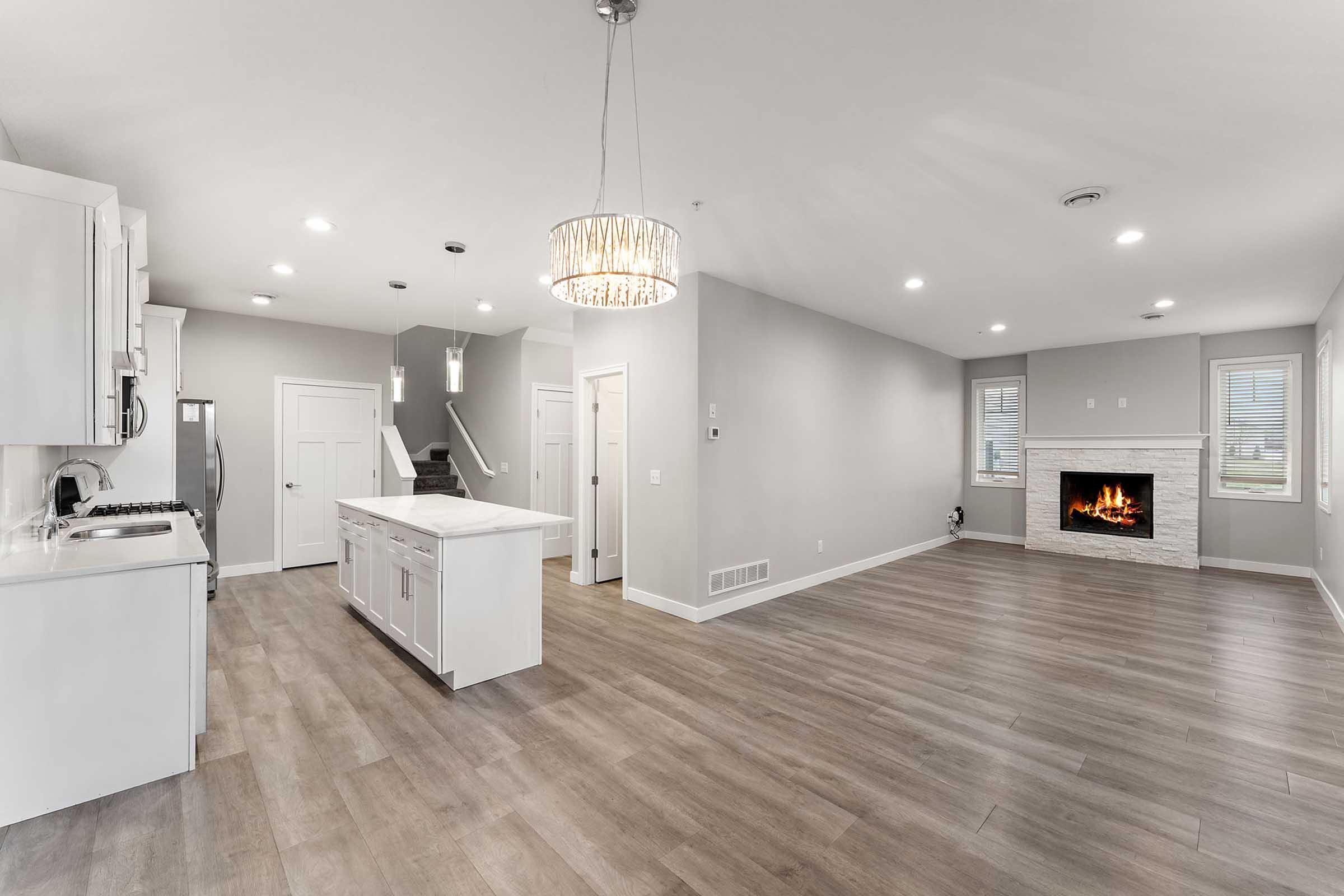 Modern open-concept living space featuring a kitchen with white cabinetry and stainless steel appliances, a dining area, and a cozy living room with a fireplace. The room is well-lit with natural light and highlights neutral tones, glossy hardwood flooring, and contemporary lighting fixtures.