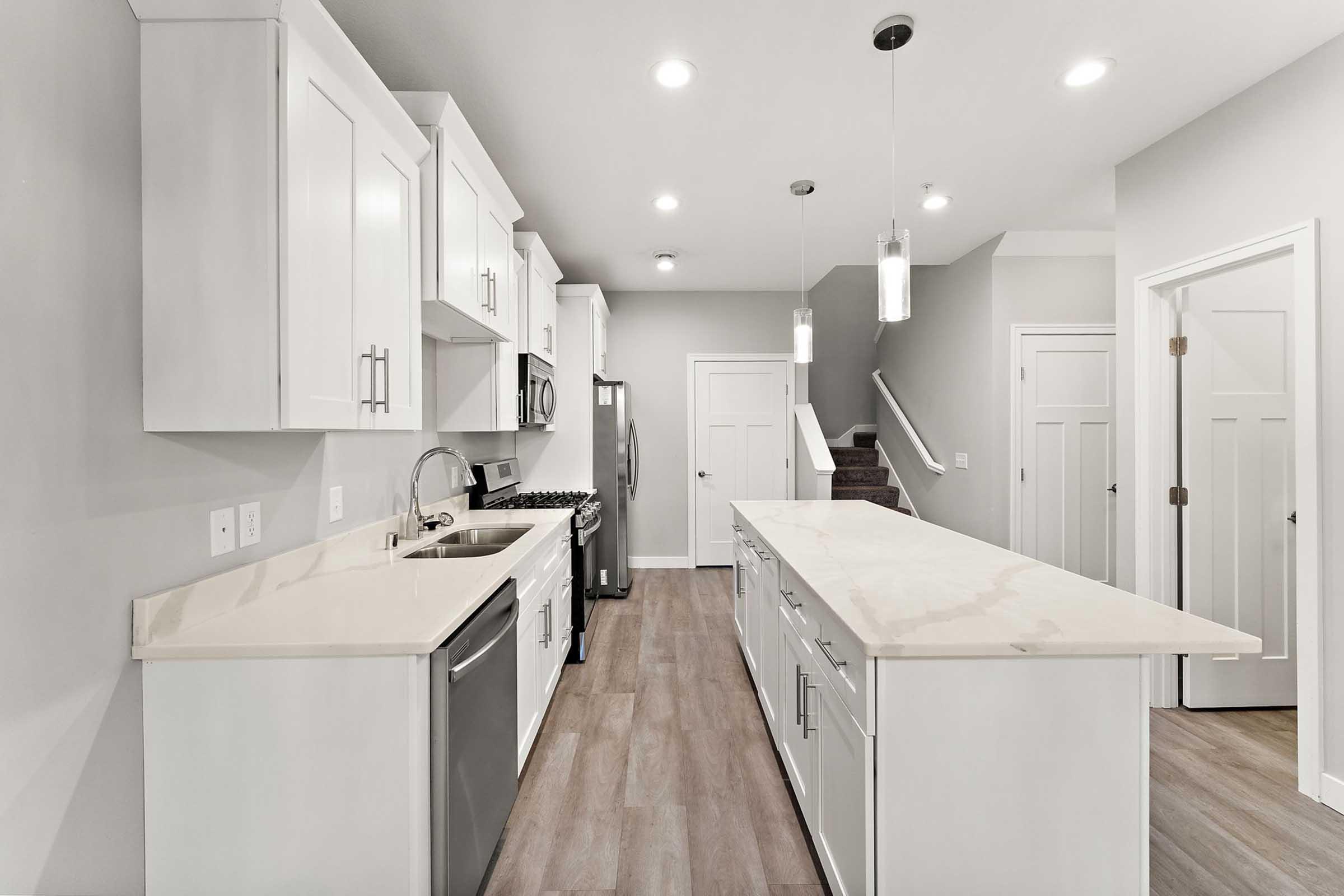 Modern kitchen featuring white cabinetry, stainless steel appliances, and a large kitchen island with a light countertop. The space has recessed lighting, a mix of open and closed storage, and a neutral color scheme with wood-like flooring. A doorway leads to a staircase in the background.