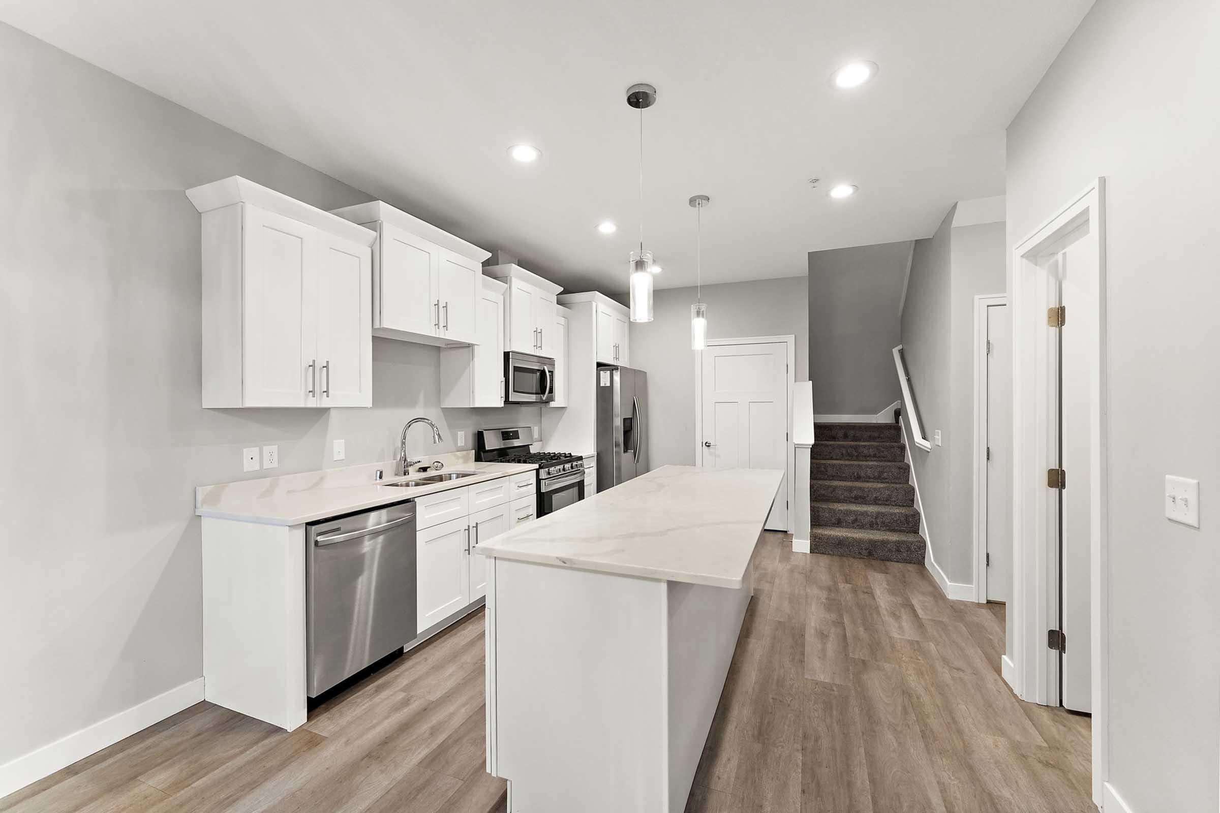 Modern kitchen featuring white cabinetry, stainless steel appliances, and a large island with a countertop. The space includes a double sink, a side-by-side refrigerator, and an open layout that connects to a stairway and living area. Bright lighting accents the neutral wall colors and wood-look flooring.