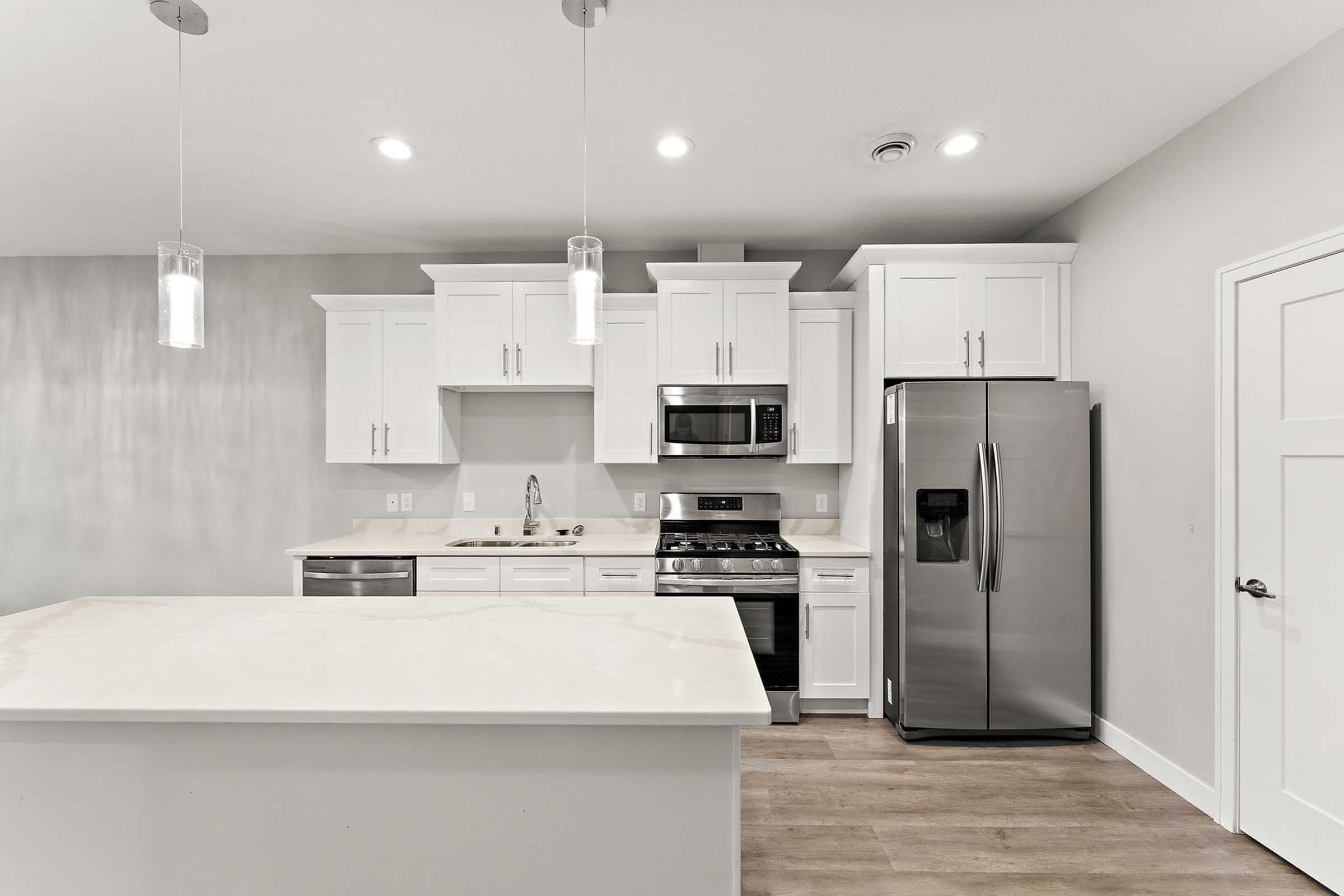 Modern kitchen featuring white cabinetry, stainless steel appliances including a refrigerator and microwave, a gas stove, and a spacious island with a light-colored countertop. The walls are painted in a neutral tone, and pendant lights hang from the ceiling, illuminating the space.