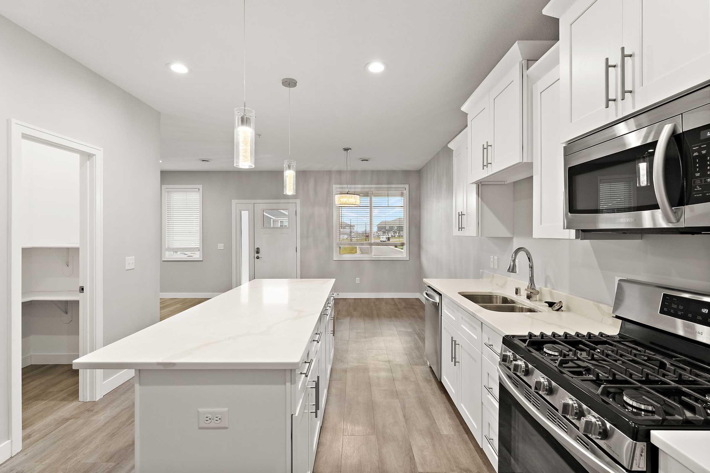 Modern kitchen with white cabinetry and stainless steel appliances. Features a spacious island with a countertop, gas stove, and sink. Brightly lit with pendant lights and large windows offering natural light. Open layout with adjacent dining area visible in the background. Warm wood-style flooring enhances the inviting atmosphere.
