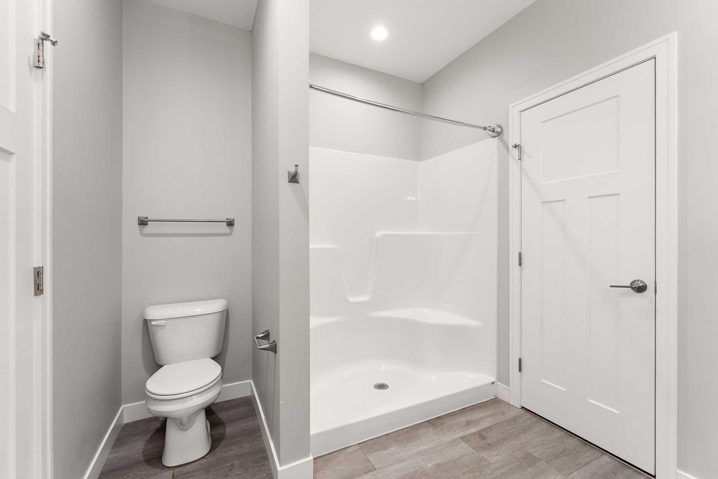 A modern bathroom featuring a white toilet and a walk-in shower with a curved glass door. The walls are painted a soft gray, and the floor has light-colored tiles. A door is visible, leading to another space, and there is a towel bar mounted on the wall near the shower. Bright overhead lighting illuminates the area.