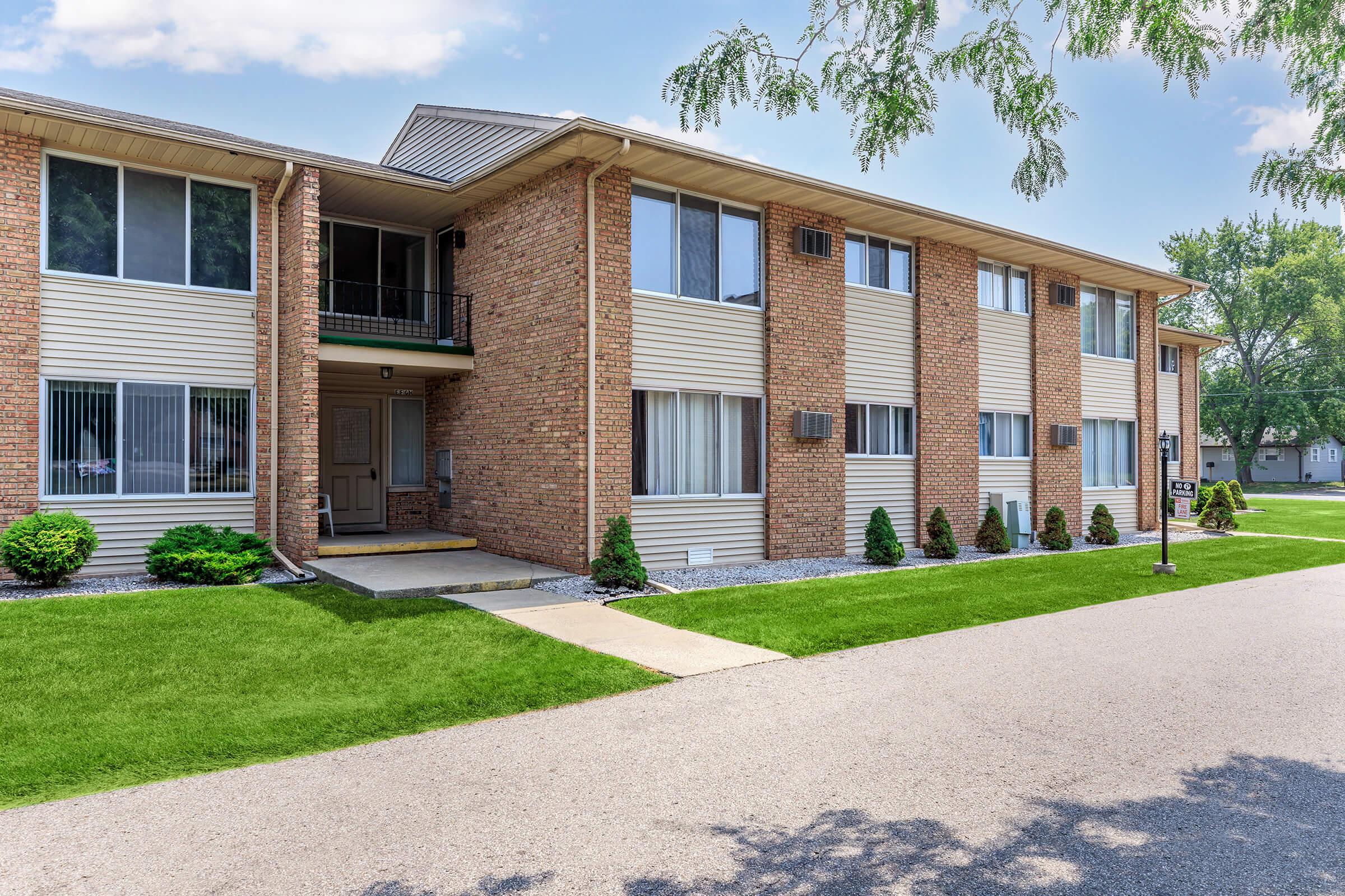 A well-maintained apartment building with a brick exterior and beige siding. The front features a walkway, manicured grass, and small shrubs. Large windows are visible on each side of the entrance, and the building is surrounded by a peaceful neighborhood setting with a clear blue sky.