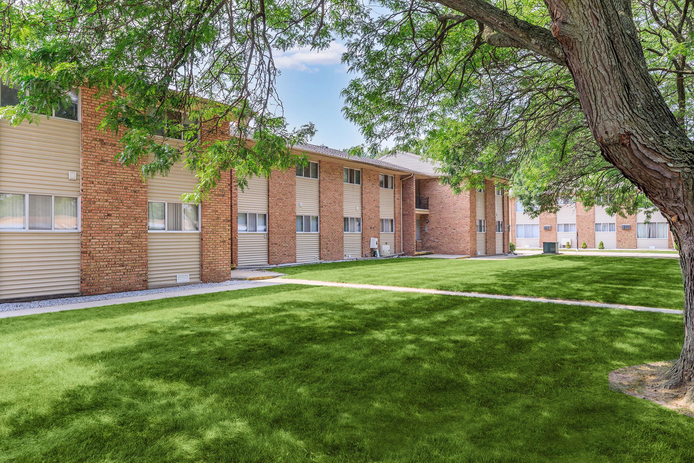 A well-maintained apartment complex with brick exterior and green lawns. The building features multiple windows, trees providing shade, and a clear blue sky. The lawn is neatly trimmed, and a path leads up to the entrance, creating an inviting residential atmosphere.