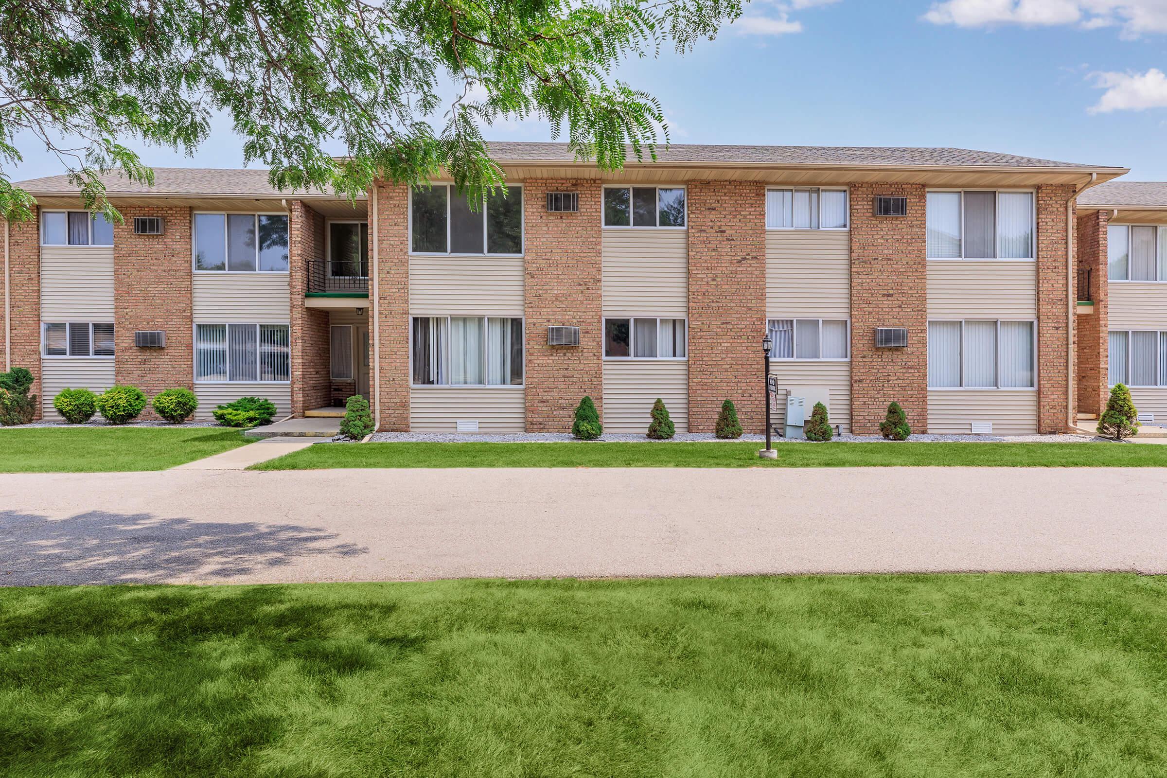 A three-story brick apartment building with a landscaped front lawn featuring small shrubs and trees. The building has multiple windows and a well-maintained pathway leading up to the entrance, under a clear blue sky.