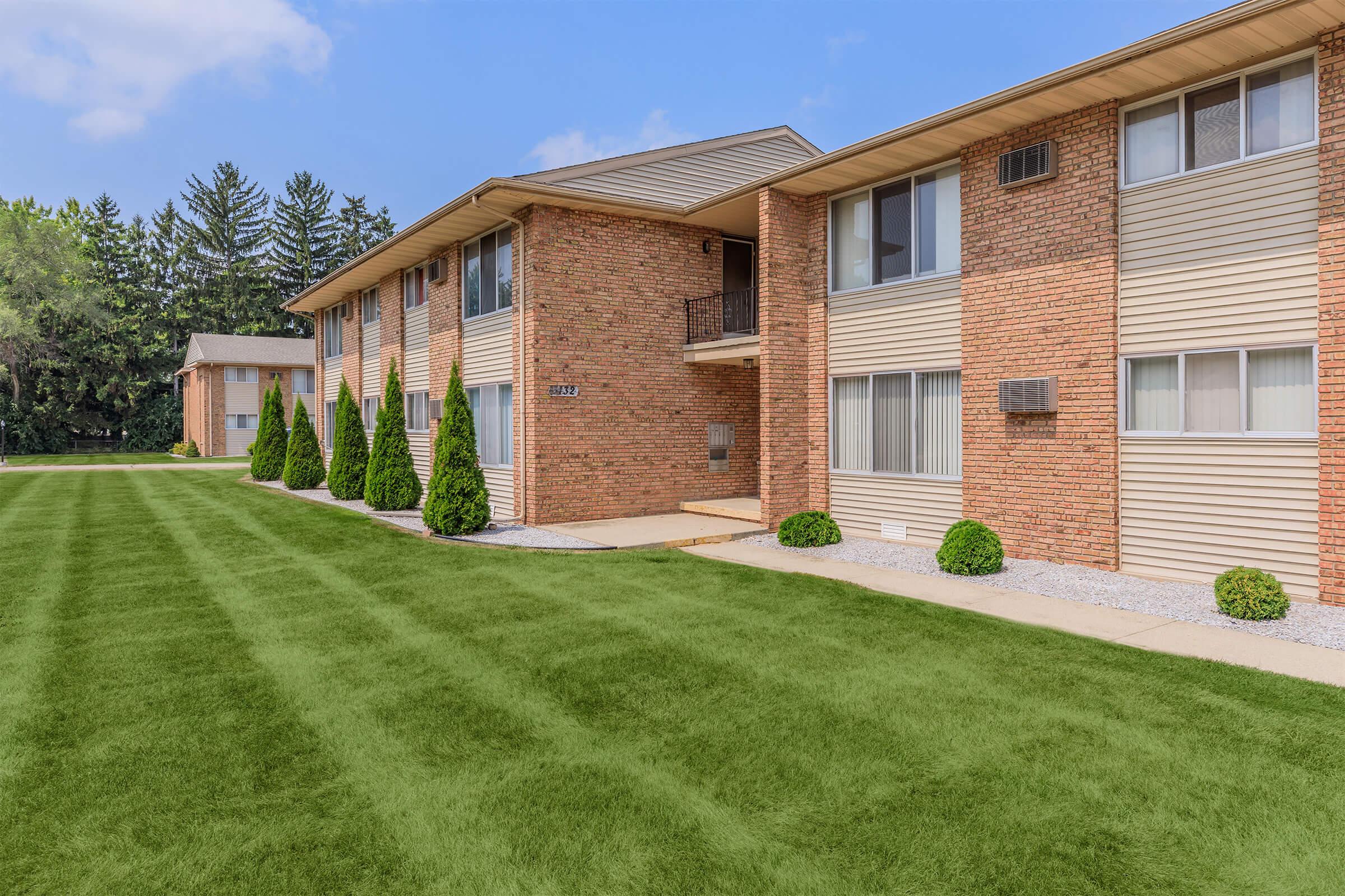 A well-maintained brick apartment building with multiple units, featuring a manicured green lawn and neatly trimmed shrubs. The sky is clear with a few clouds, and the surrounding area includes trees, creating a pleasant residential environment.
