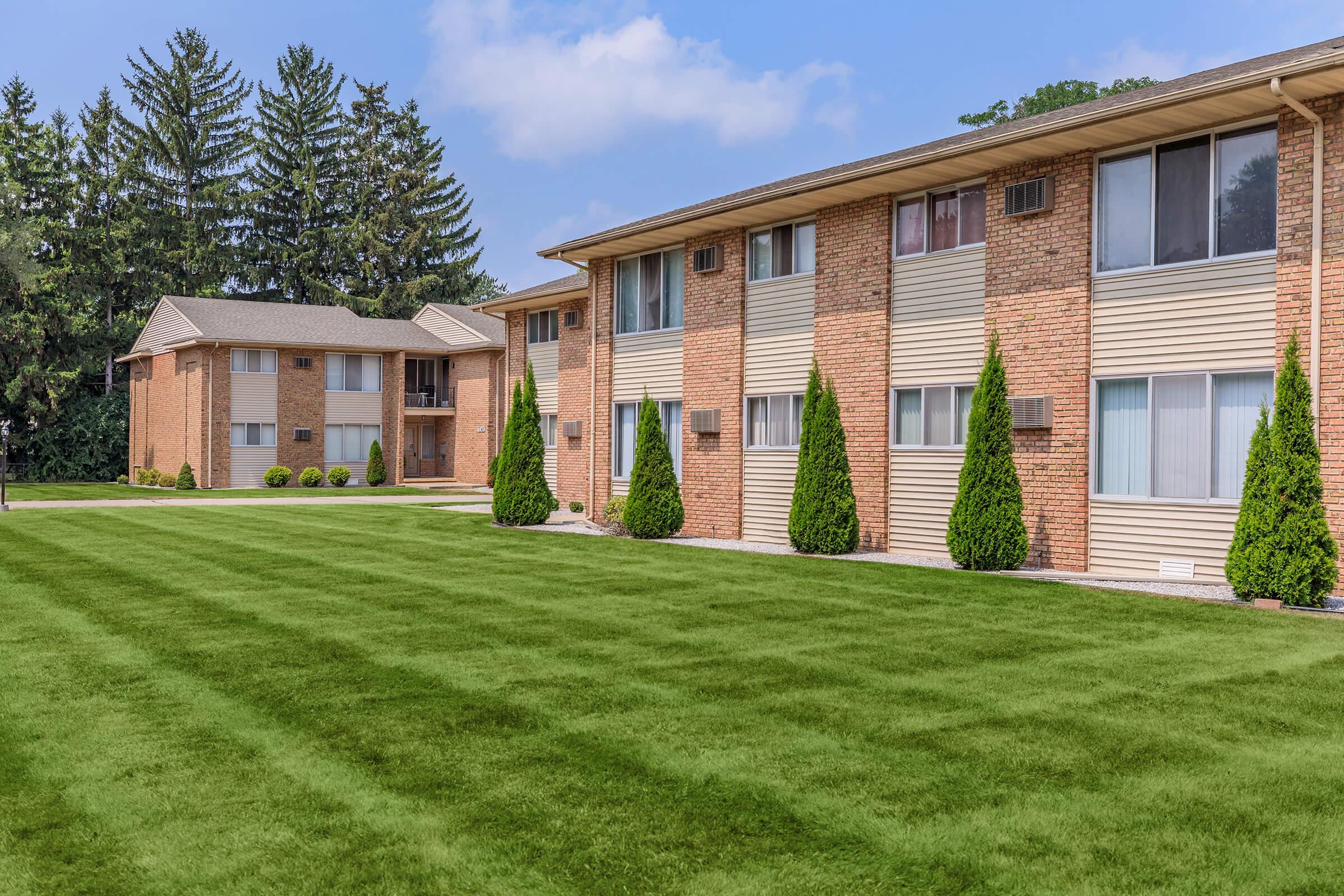 A well-maintained apartment complex featuring two buildings made of brick and siding. The foreground has a lush green lawn with neatly trimmed bushes, while tall trees provide a natural backdrop. The sky is partly cloudy, creating a pleasant and inviting atmosphere.
