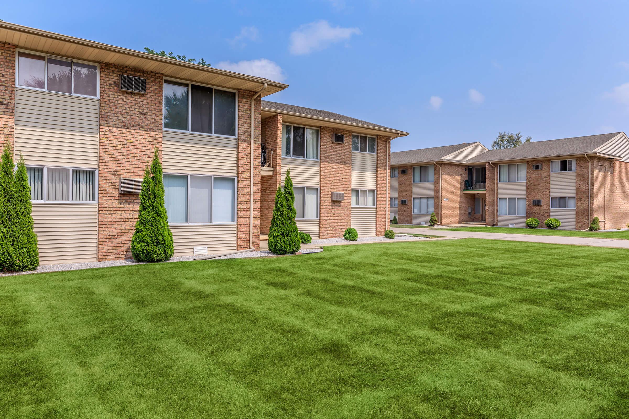 Two brick apartment buildings with multiple windows are located side by side. A well-maintained green lawn with neatly trimmed bushes in front of the buildings creates a clean, inviting atmosphere. The sky is clear and blue, enhancing the overall pleasant appearance of the scene.