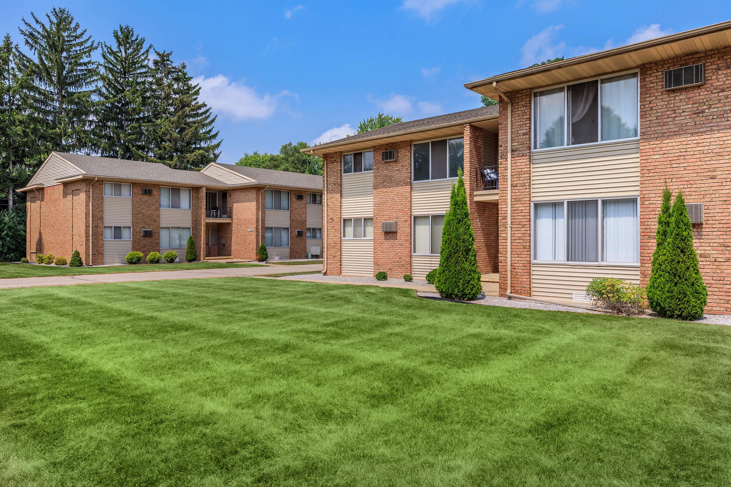 A well-maintained residential apartment complex featuring two brick buildings. The foreground shows a neatly manicured green lawn, while the background has trees lining the property and a clear blue sky above. The buildings have large windows and are surrounded by small bushes.