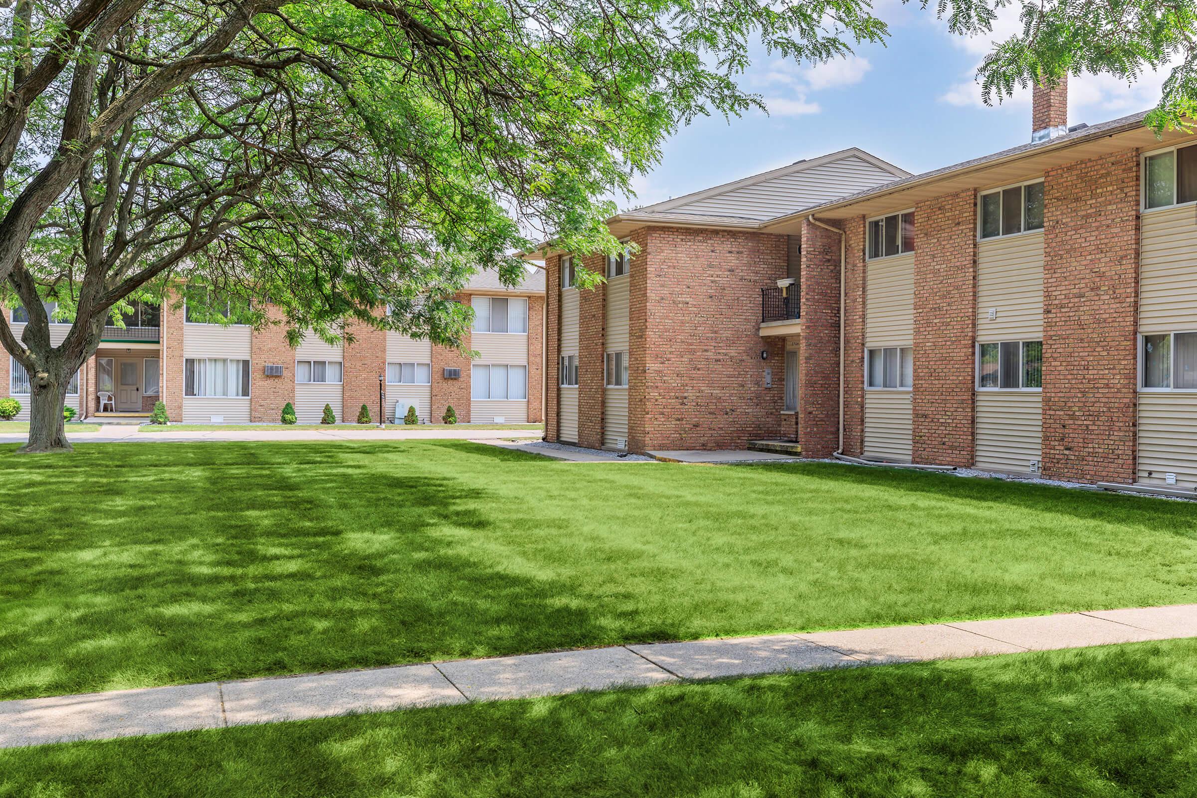 A well-kept apartment complex featuring brick and siding exteriors, surrounded by lush green grass and shaded by trees. The landscape includes a wide walkway leading through the grassy area, with multiple residential units visible in the background.