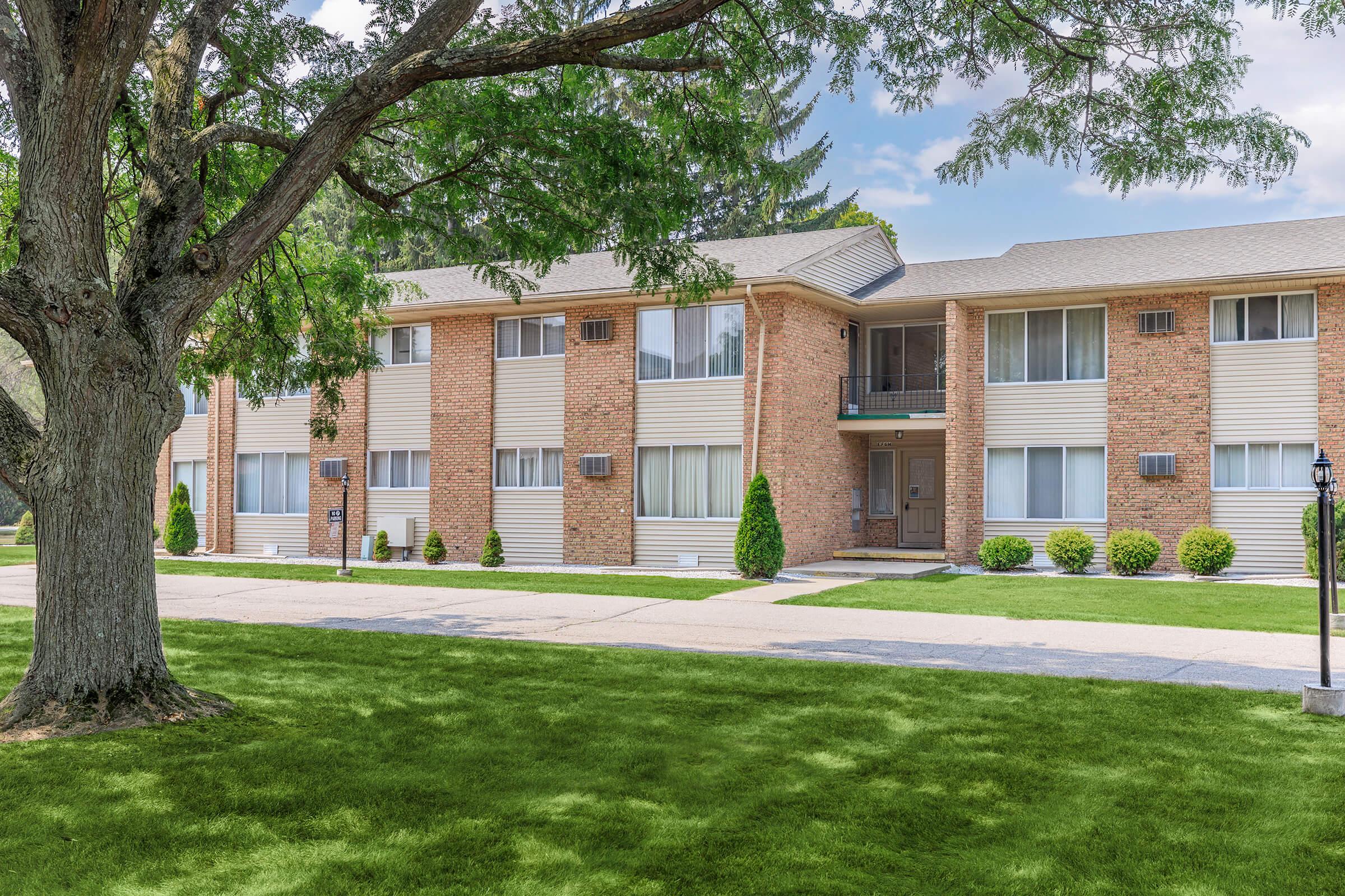 A two-story brick apartment building with large windows and green lawn. A tree is in the foreground, providing shade. The building features neatly trimmed shrubs and a paved pathway leading to the entrance. The scene is bright and inviting, set against a clear sky.