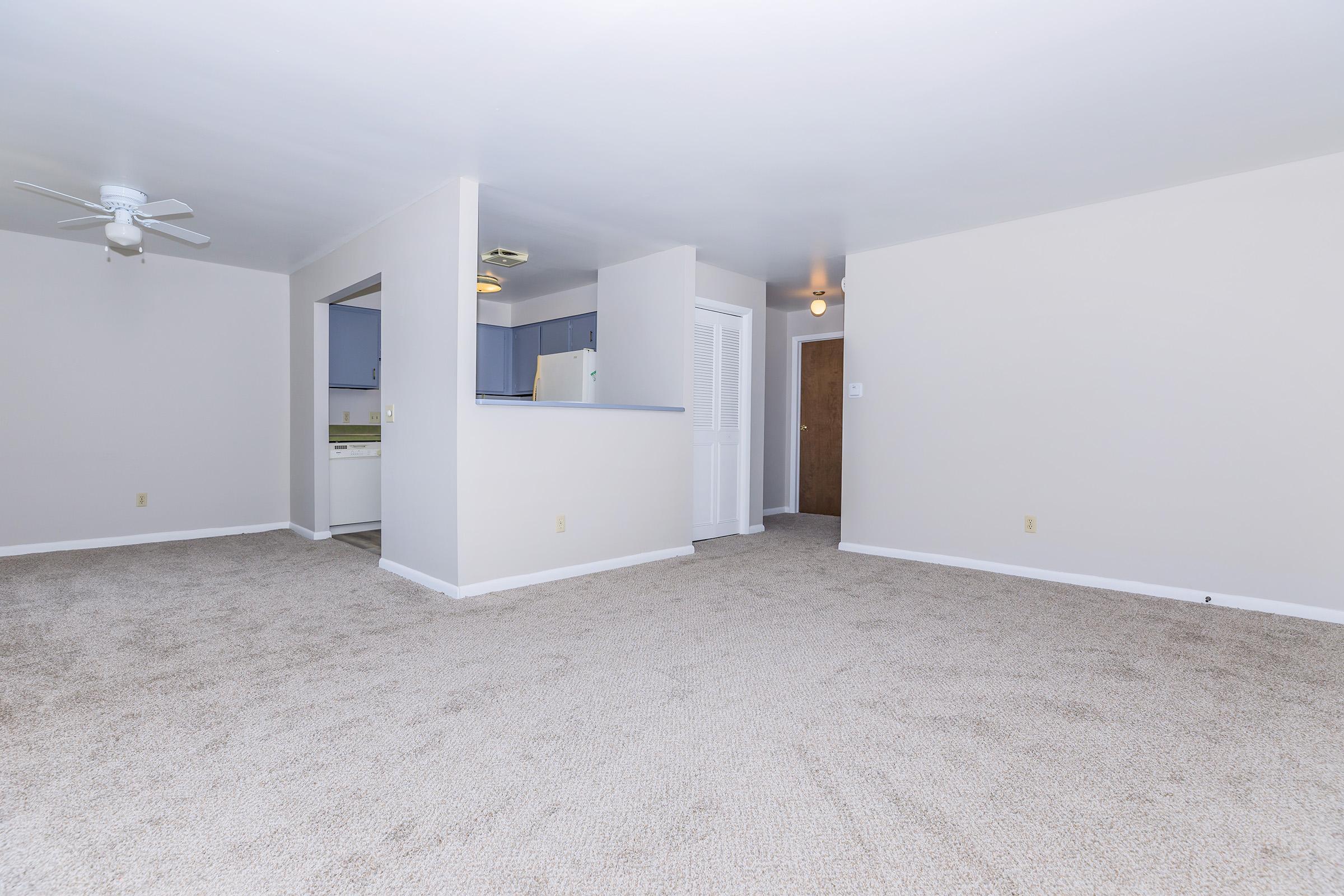 A clean, empty living room with light-colored walls and carpet. A ceiling fan is visible, along with a doorway leading to another room. There is an open kitchen area in the background, showcasing cabinets and appliances. The space is well-lit and has a neutral aesthetic.