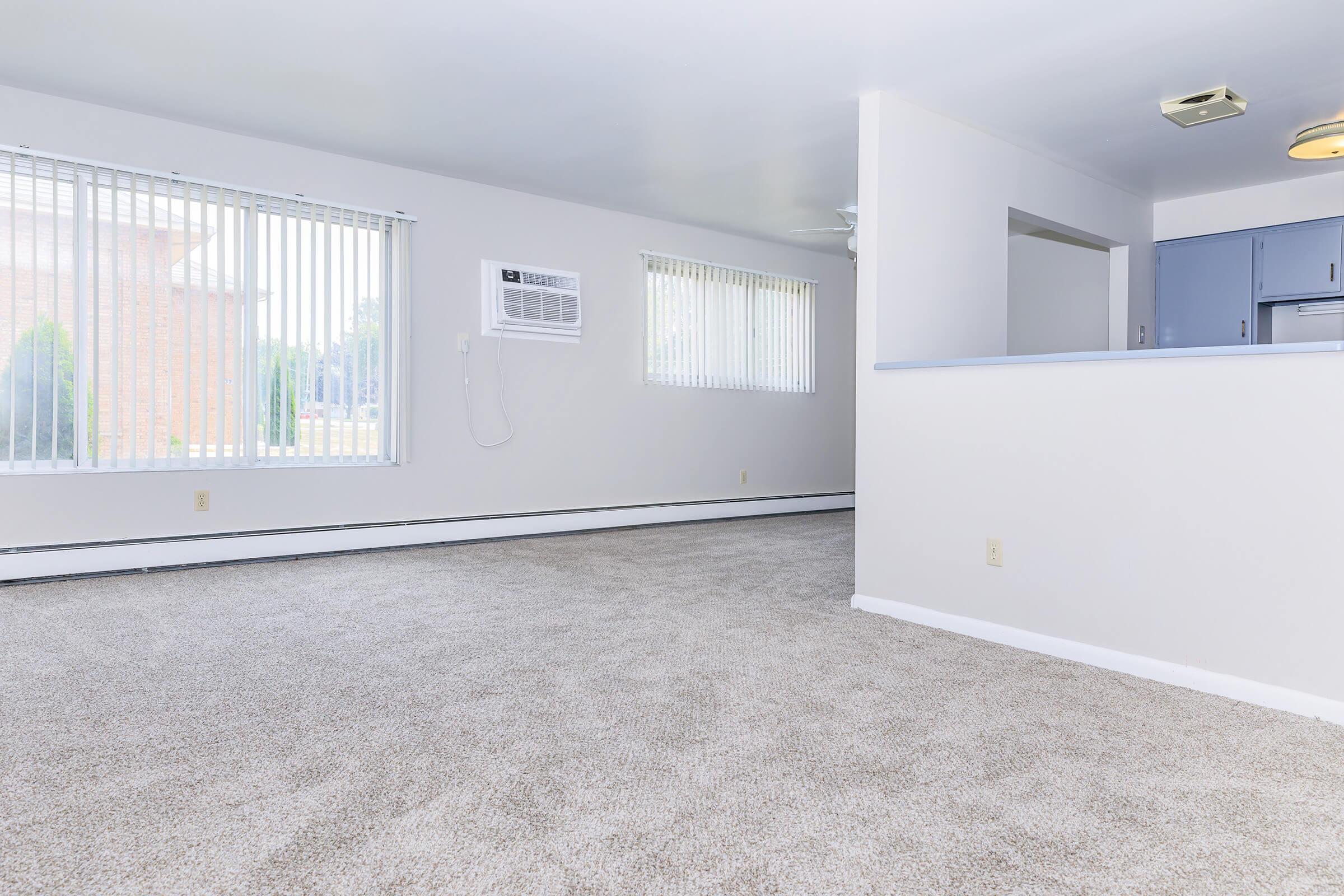 A spacious, light-filled room with neutral-colored walls and carpet. Two large windows with blinds allow natural light to enter. An air conditioning unit is installed on one wall, and there's a small kitchen area visible in the background. The space appears clean and inviting, suitable for living or entertaining.