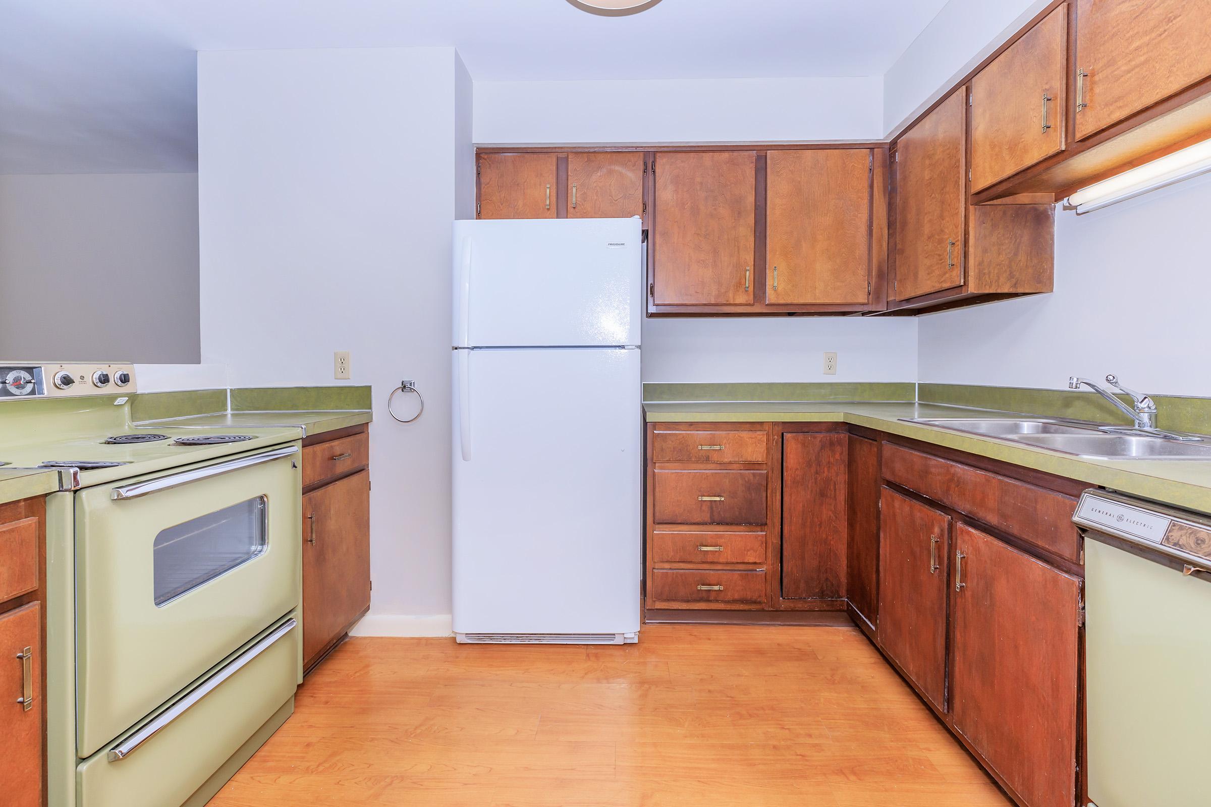 Bright kitchen featuring wooden cabinets and green countertops. Includes a white refrigerator, an oven with a mint green finish, and a stainless steel sink. The floor is a light wood laminate, creating a warm and inviting atmosphere. A window is absent, making the space feel simple and functional.