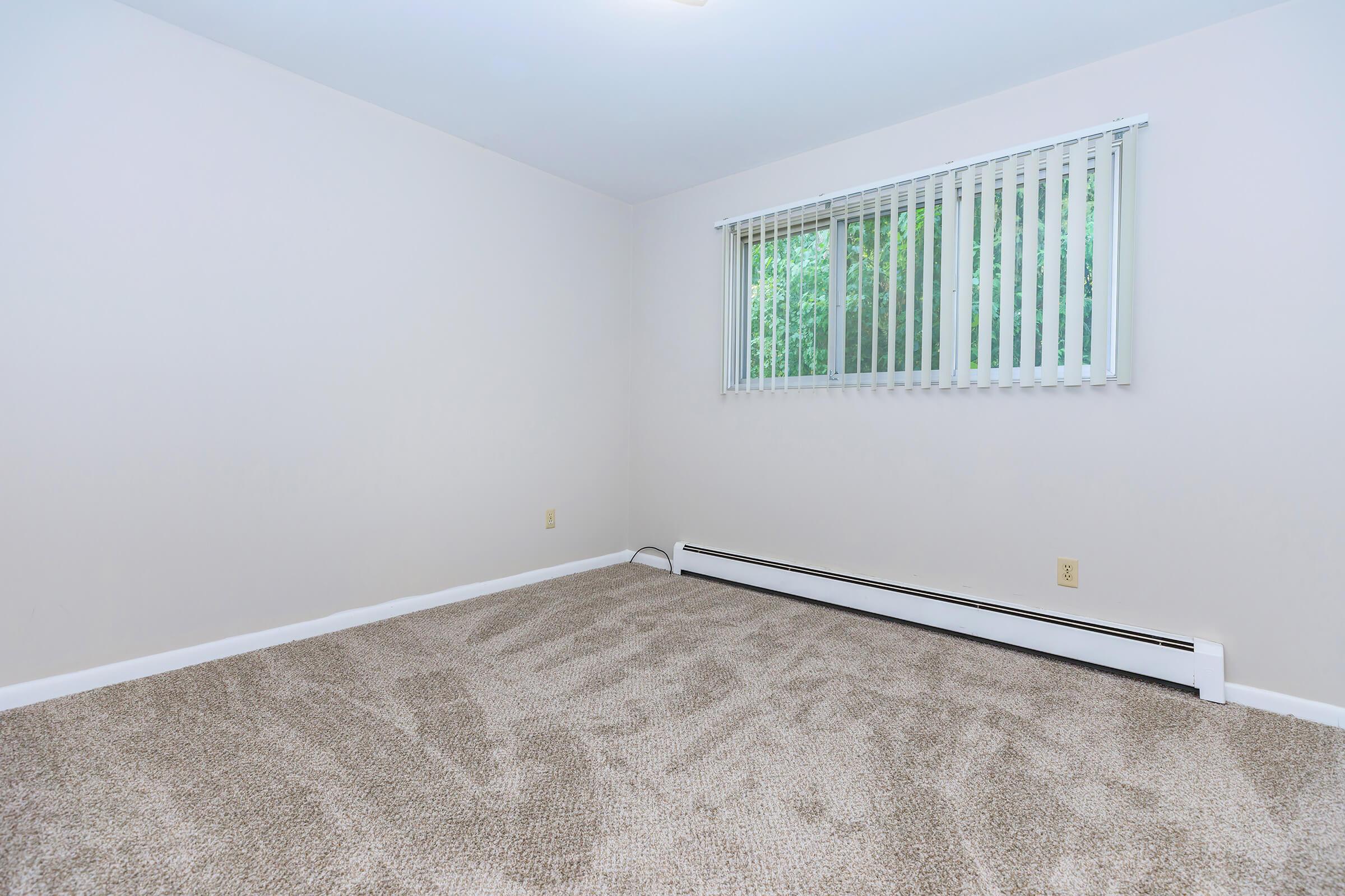 A vacant room with beige walls and light brown carpet. The space has a window with vertical blinds allowing natural light to enter. There are no furnishings or decorations, creating a minimalist and open atmosphere.