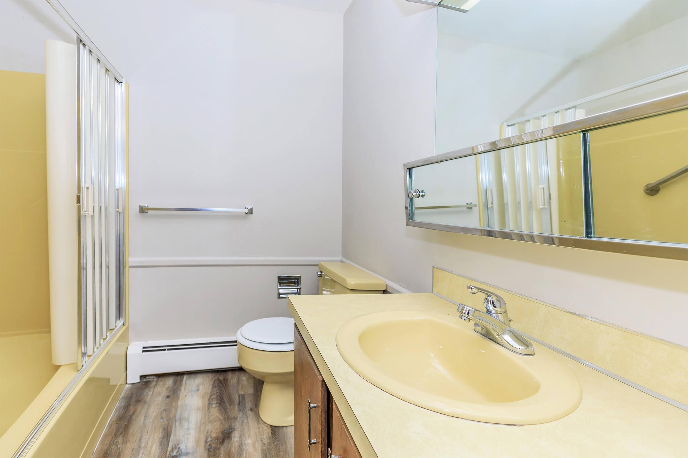 A bathroom featuring a yellow bathtub and toilet, a beige countertop with a sink, a large mirror above the sink, and grab bars on the wall. The flooring is wood-like, and the overall decor is simple and functional. The walls are painted light colors, enhancing the space's brightness.