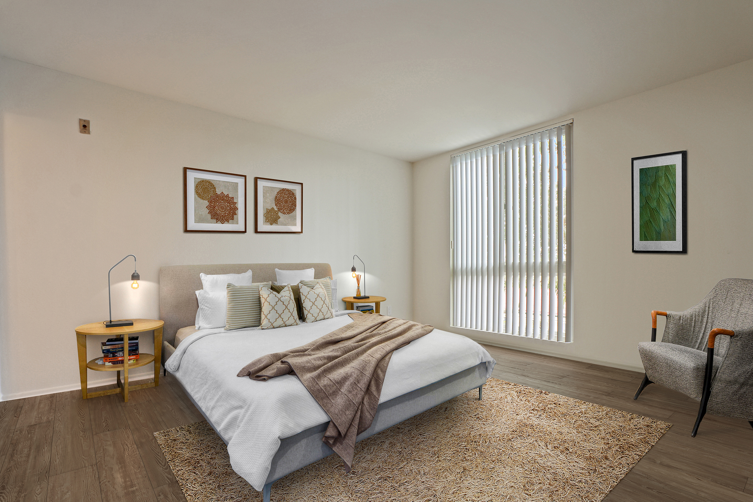 A cozy bedroom featuring a neatly made bed with white linens and decorative pillows. There are two bedside tables with lamps and books. A soft rug covers the floor, while large windows with vertical blinds allow natural light. An armchair adds to the welcoming atmosphere, complemented by framed artwork on the walls.