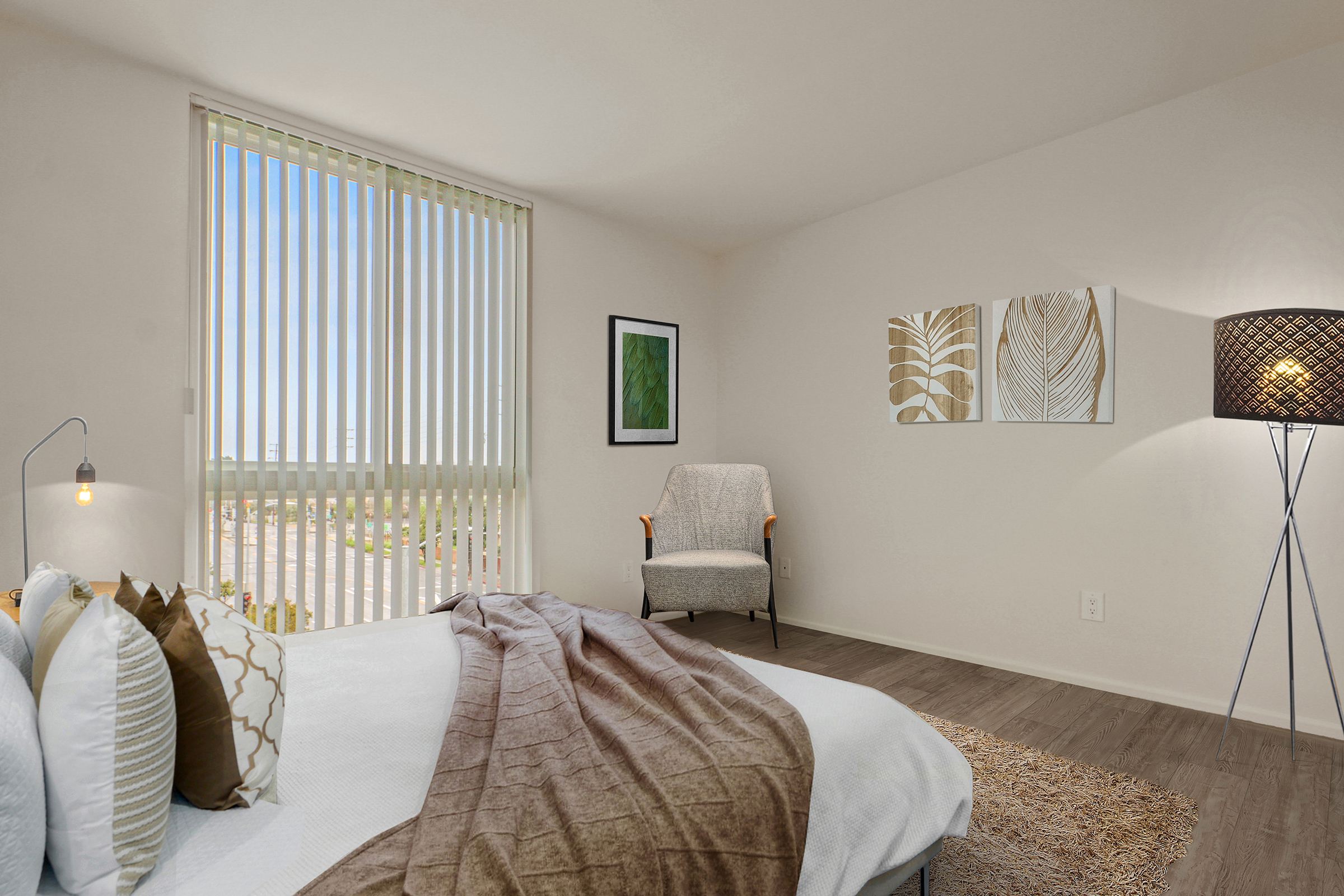 A cozy and modern bedroom with a large window showing blue skies. The bed is adorned with decorative pillows and a warm throw. There's a stylish chair in the corner, a floor lamp, and framed leaf artwork on the wall, creating a welcoming and serene atmosphere.