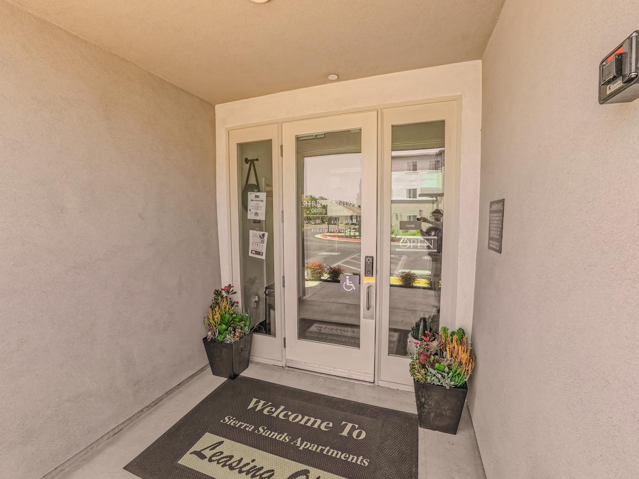 Entrance to Sierra Sands Apartments featuring double glass doors with a welcoming mat that reads "Welcome To" and decorated with potted plants on either side. The area is well-lit and includes signage adjacent to the door.
