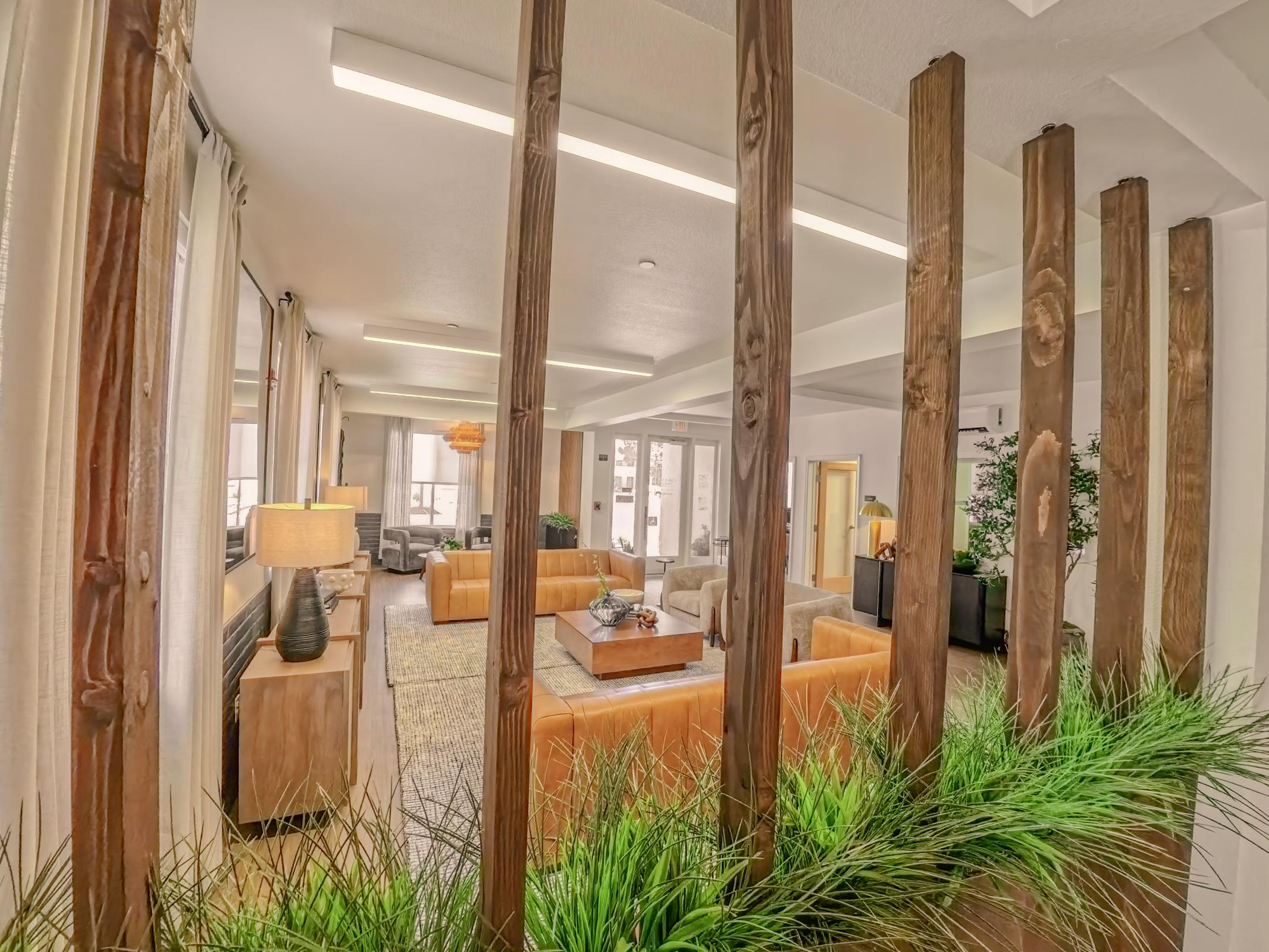 A bright, modern living space featuring two orange sofas, a coffee table, and large windows. There are wooden beam dividers and green plants in the foreground. Soft lighting creates a warm atmosphere, with a lamp and decorative elements enhancing the inviting design.