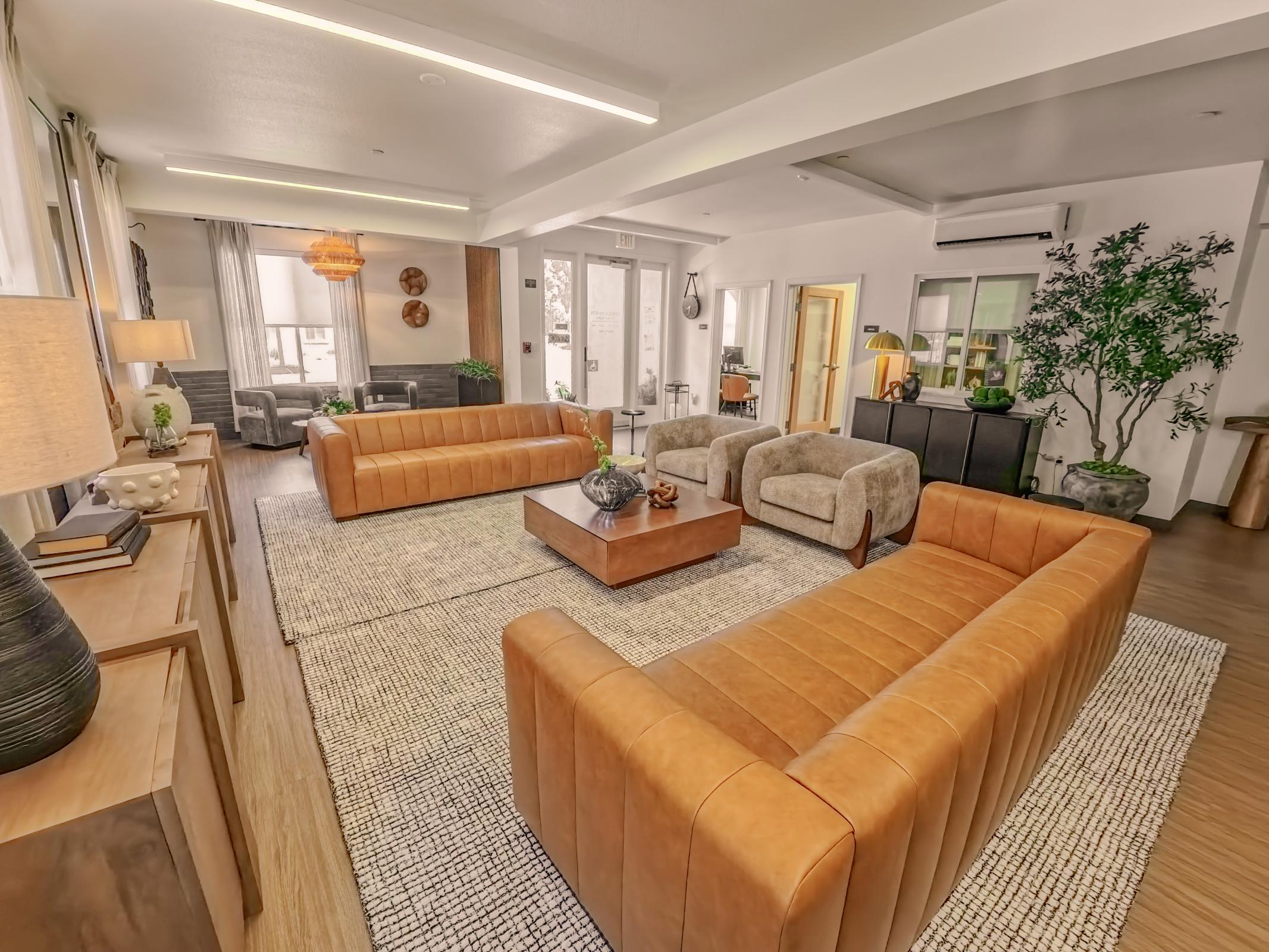 A spacious living room featuring two brown leather sofas, two beige armchairs, a wooden coffee table with a decorative vase, and a textured rug. Large windows allow natural light to illuminate the space, which is accented by houseplants and modern decor elements.