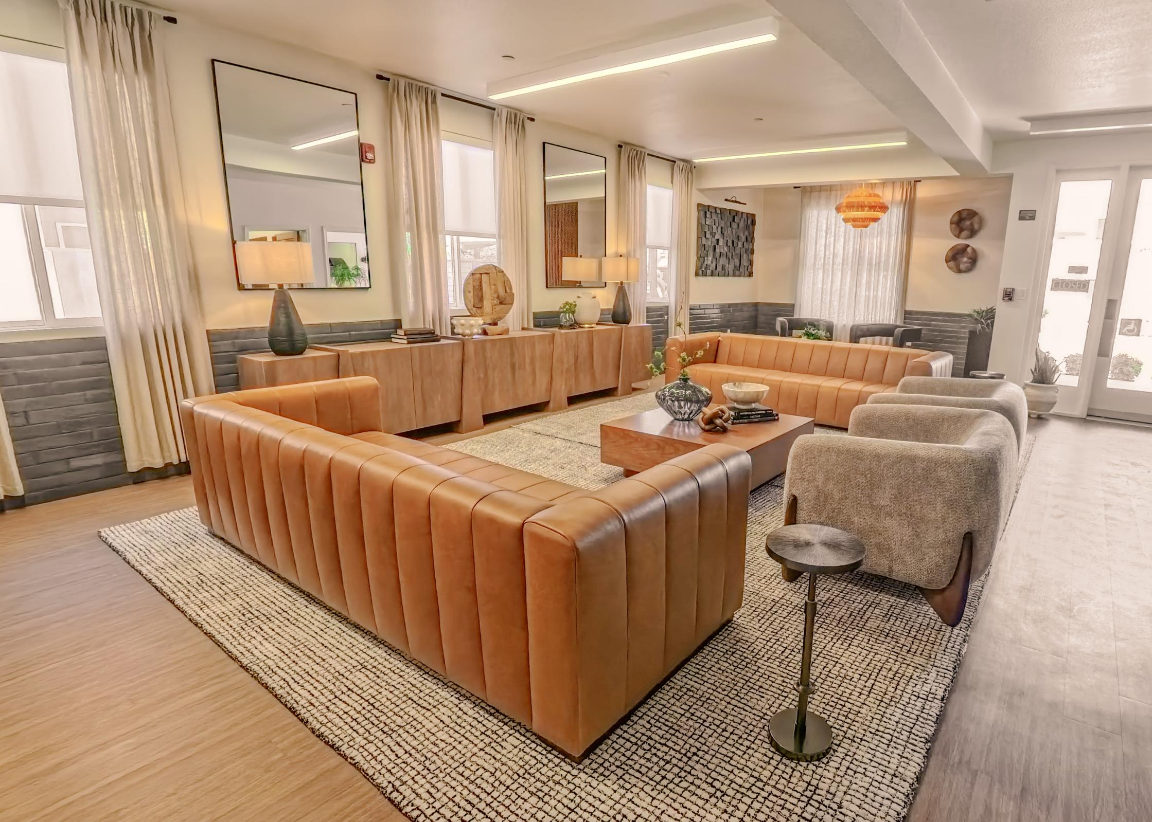 A spacious modern living room featuring a large, brown leather sectional sofa, a wooden coffee table, and stylish decor. Natural light streams through large windows, complemented by beige curtains. The space is accented with a patterned area rug and contemporary furnishings, creating a warm and inviting atmosphere.